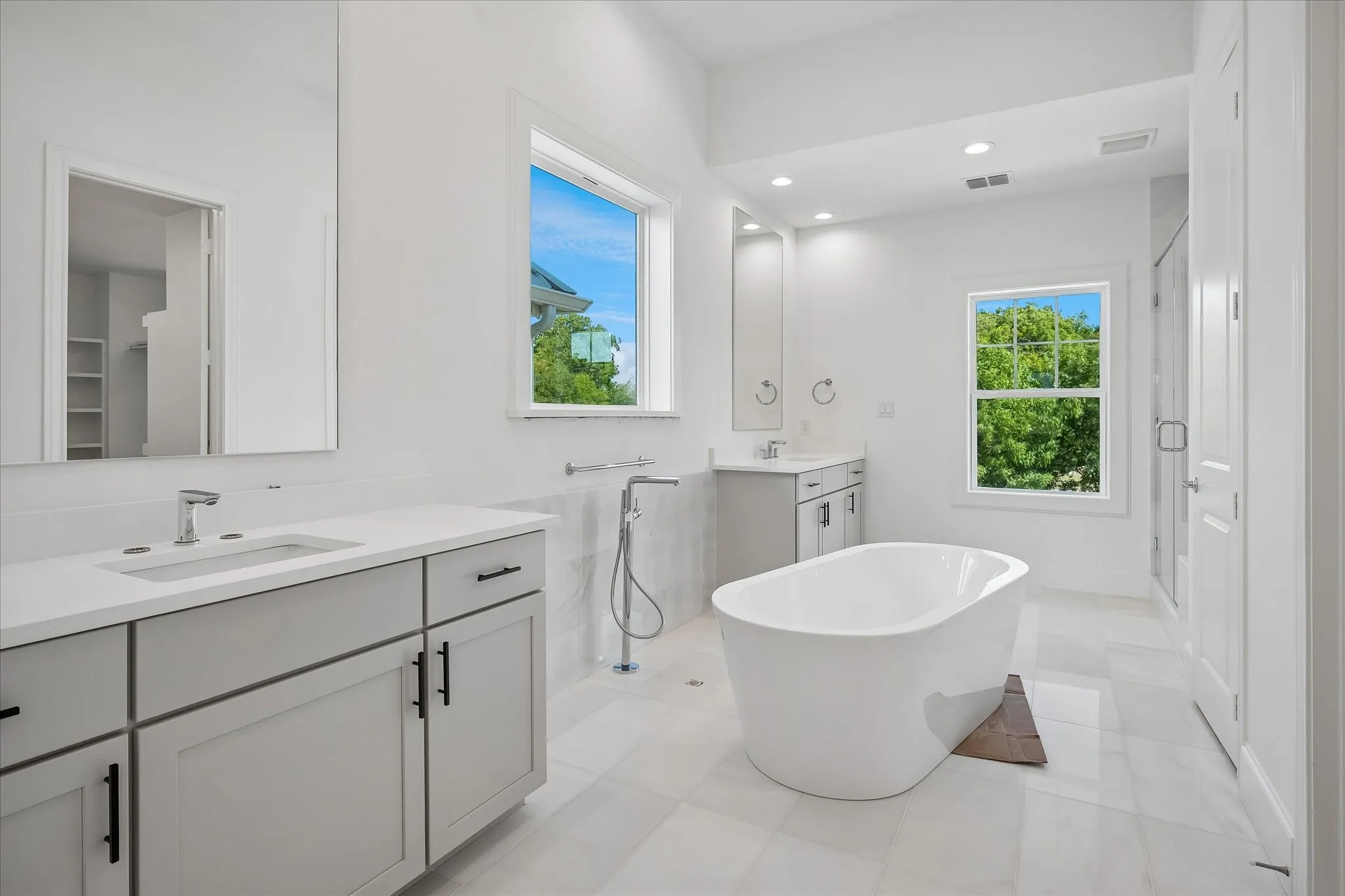 Spa-inspired primary bathroom featuring a freestanding soaking tub, dual vanity with quartz countertops and matte black hardware, walk-in shower with glass enclosure and rainfall showerhead, polished tile flooring, and abundant natural light from multiple windows