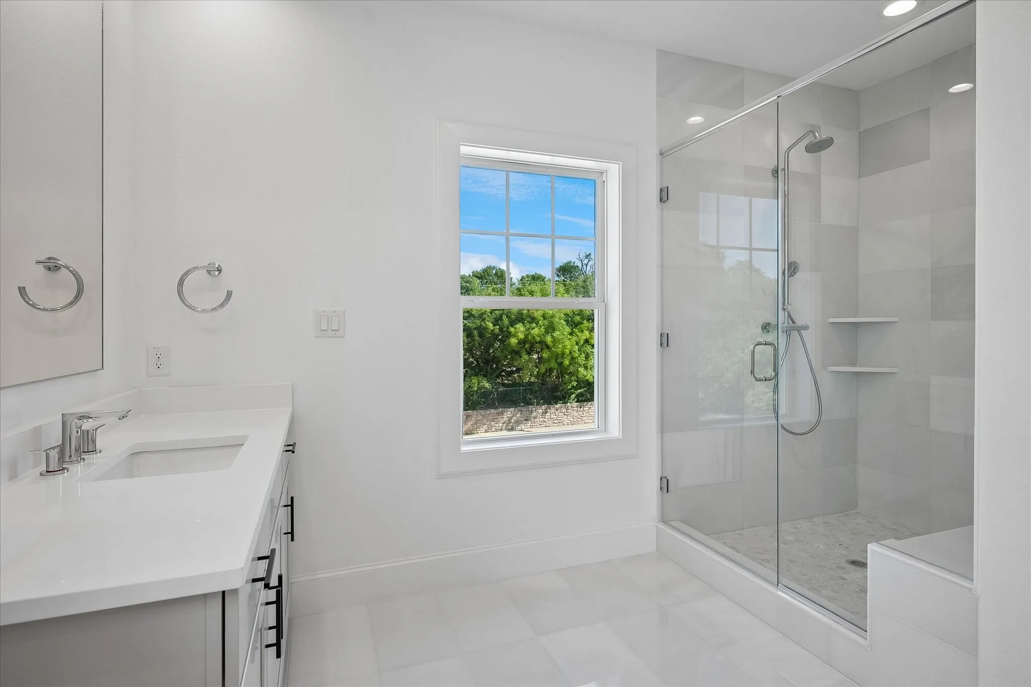 Spa-inspired primary bathroom featuring a frameless glass-enclosed shower with polished chrome fixtures, floating white vanity with rectangular vessel sink and chrome faucet, white porcelain tile flooring, recessed ceiling lighting, and a large casement window framing natural landscaping views
