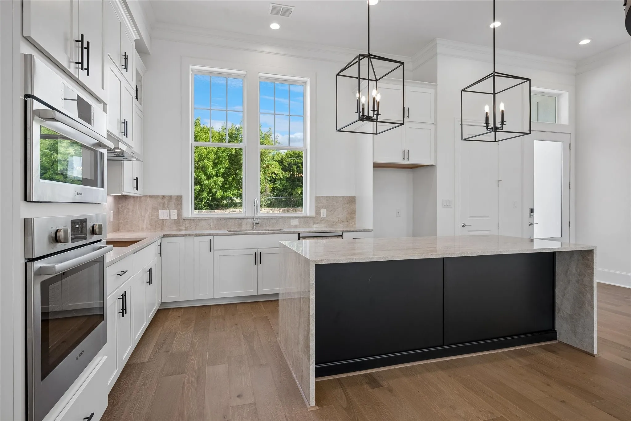 Modern kitchen featuring white cabinetry with hardware accents, stainless steel built-in appliances including a wall oven, quartz countertops, a substantial black-finished island with waterfall edge, wood-finish flooring throughout, and geometric pendant lighting