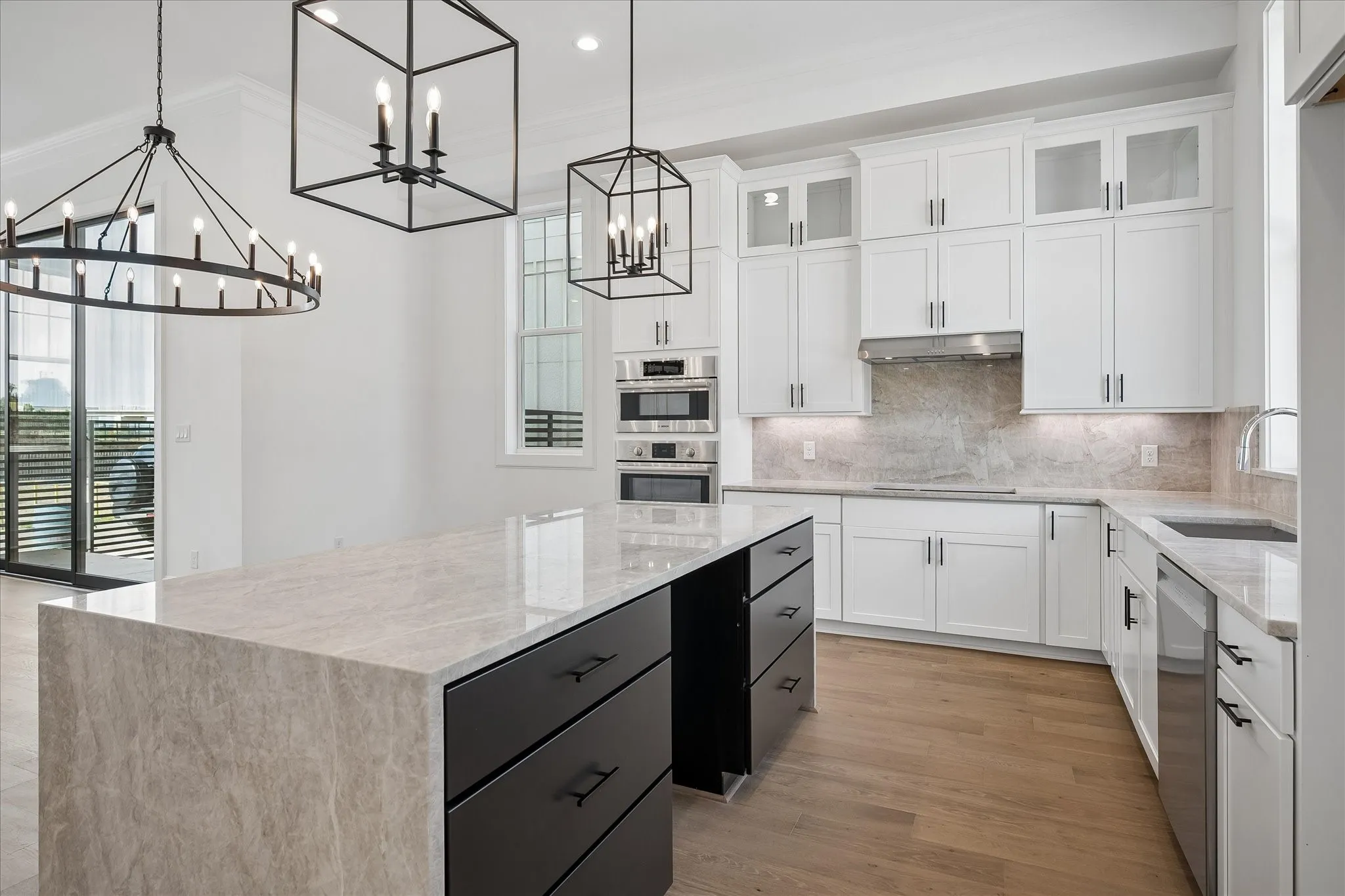 Chef's kitchen featuring white cabinetry with glass-front uppers, quartz countertops, a large waterfall-edge island with black base cabinetry, stainless steel double wall ovens, and warm wood-finish flooring