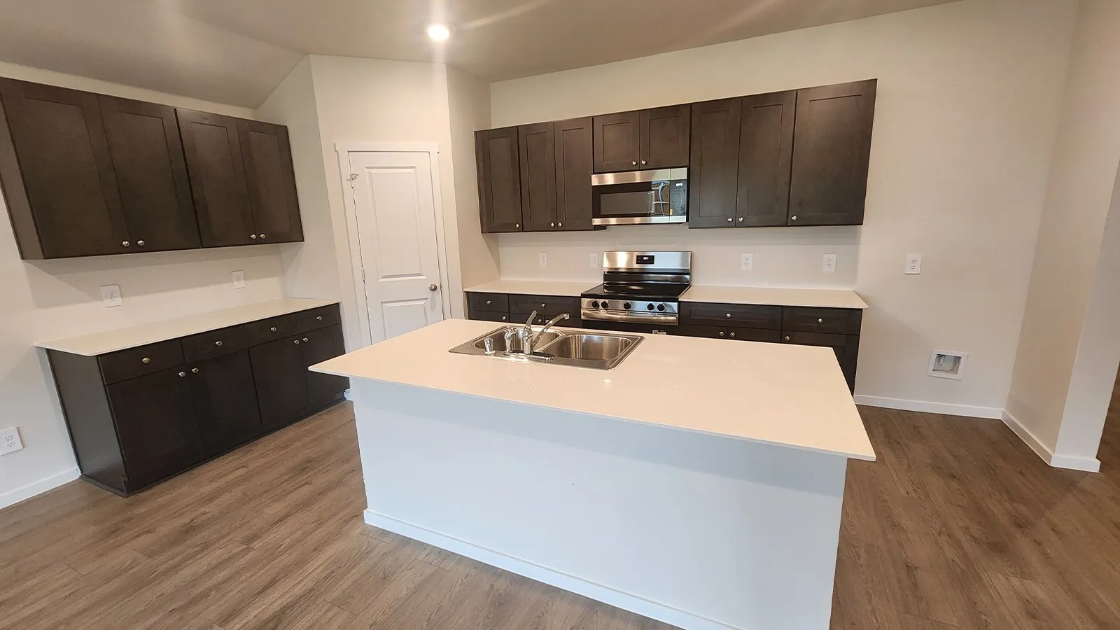 Modern kitchen featuring an island with an undermount double basin sink, dark wood-finish cabinetry, light countertops, stainless steel appliances, and wood-finish flooring