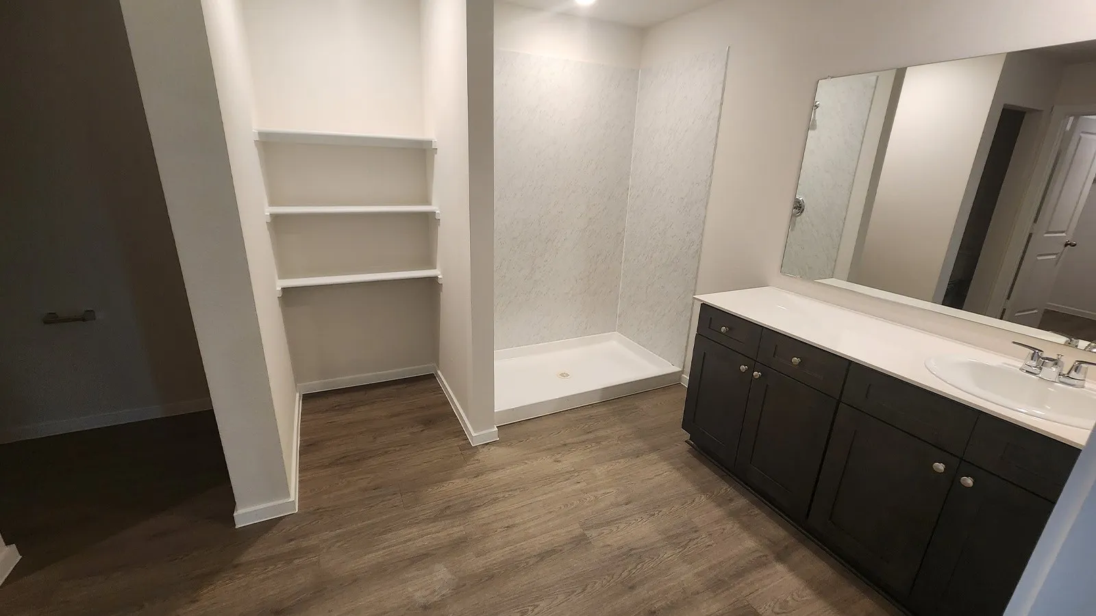 Bathroom featuring a wood-finish floor, a dark wood-finish vanity with a white countertop and integrated sink, a large wall-mounted mirror, a walk-in shower with textured wall panels, and built-in shelving