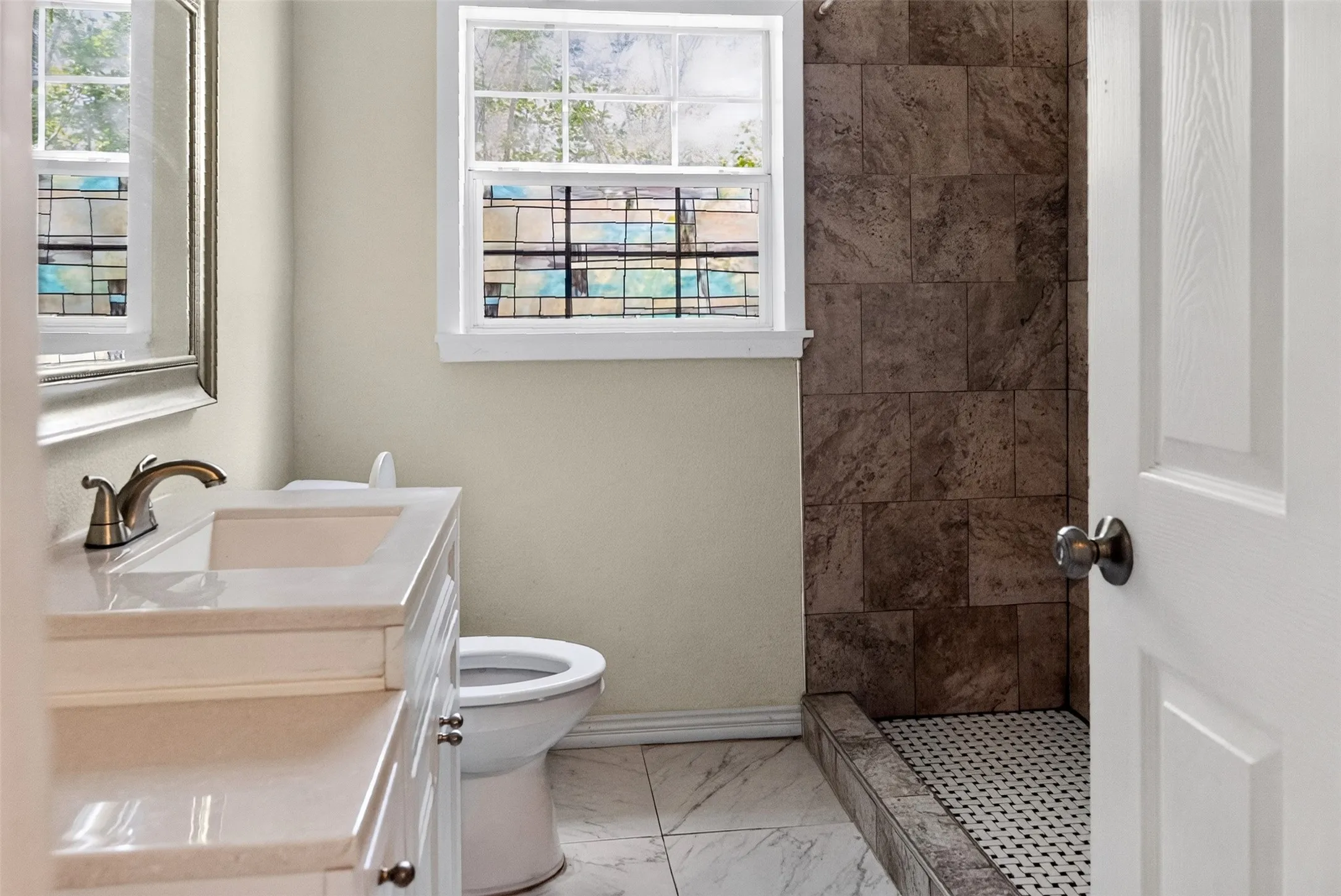 Bathroom featuring a white vanity with a light-toned countertop, a shower with dark-toned tile walls and a mosaic tile floor, and a white framed window with decorative glass