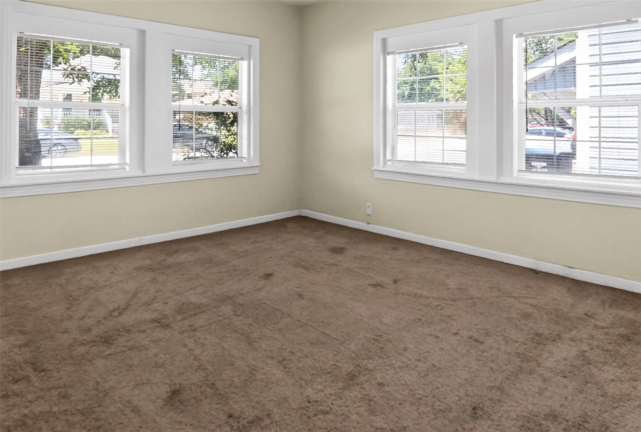 Interior room featuring three windows with white blinds, light beige painted walls, white baseboards, and brown carpet flooring