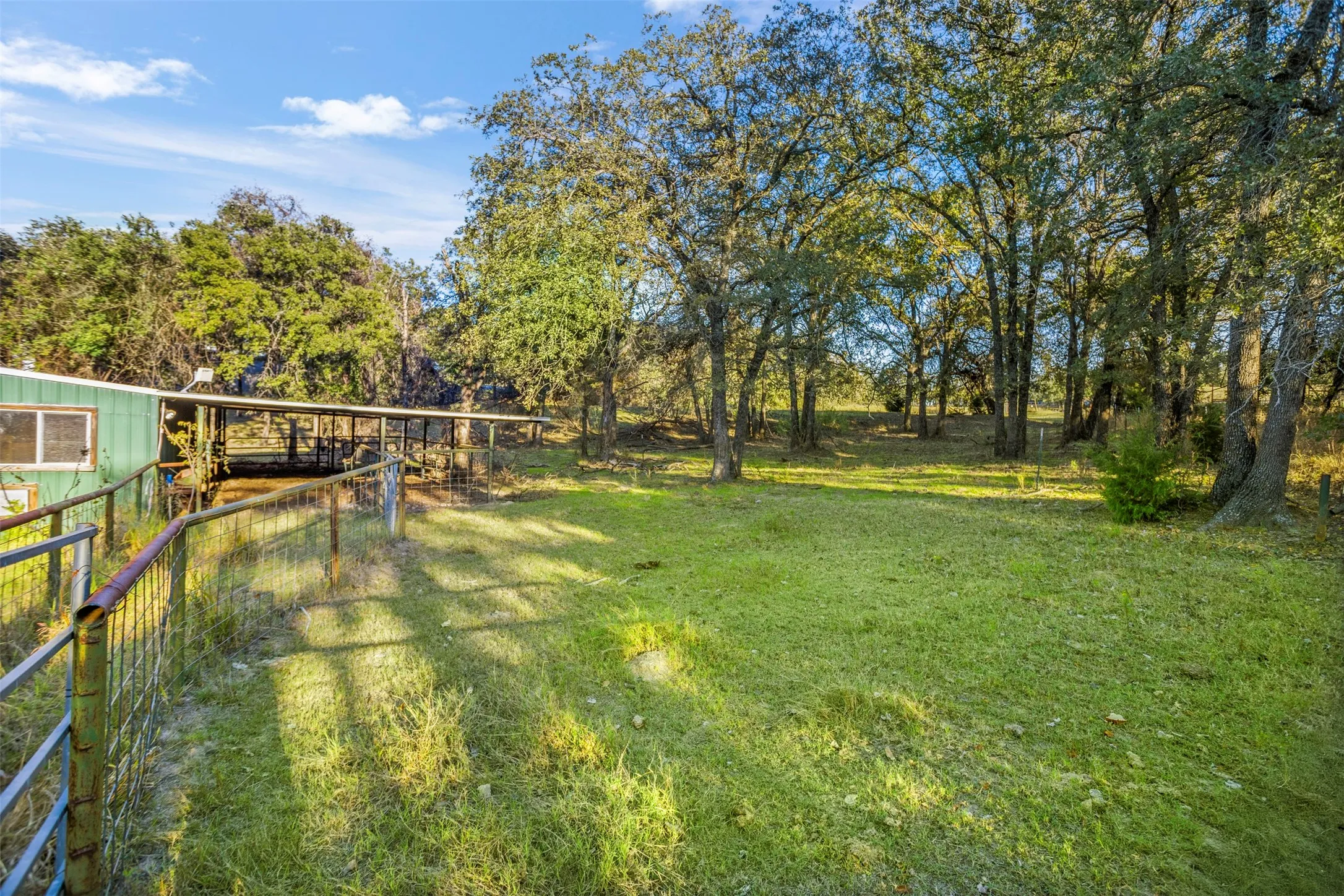 Expansive greenspace featuring mature trees, a fenced enclosure, and a covered structure with an extended overhang