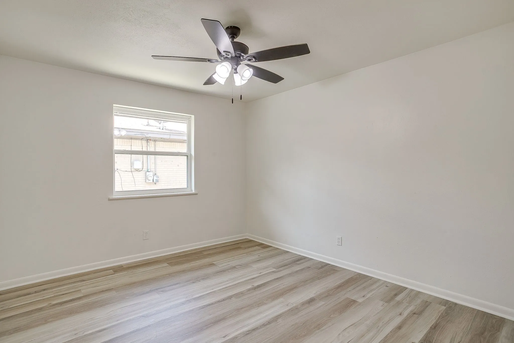 Bright room featuring light wood-finish flooring, white walls, a single window, and a ceiling fan with integrated lighting