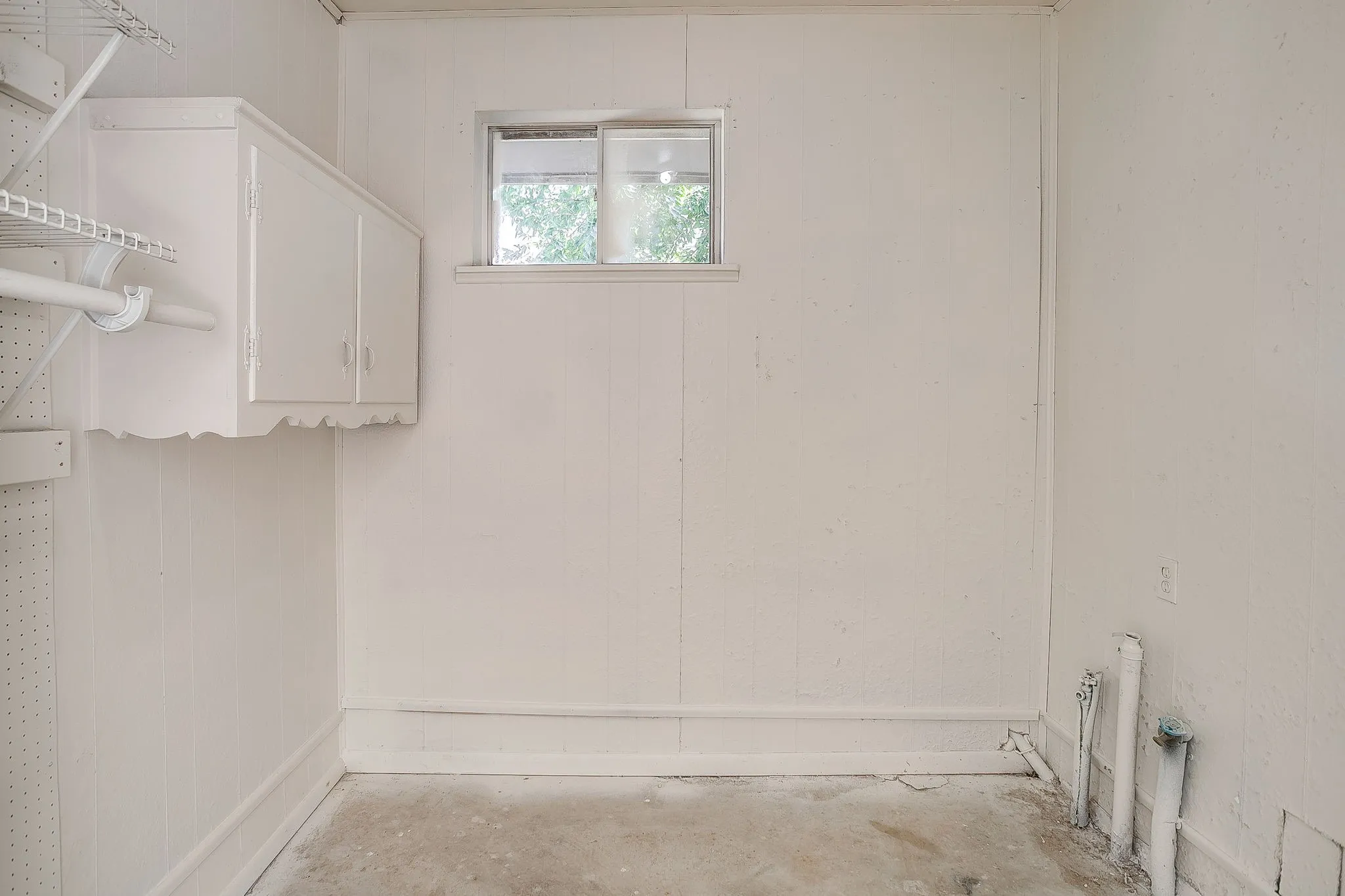 Utility room featuring white paneling, a single window, built-in white cabinetry, metal shelving, and concrete flooring