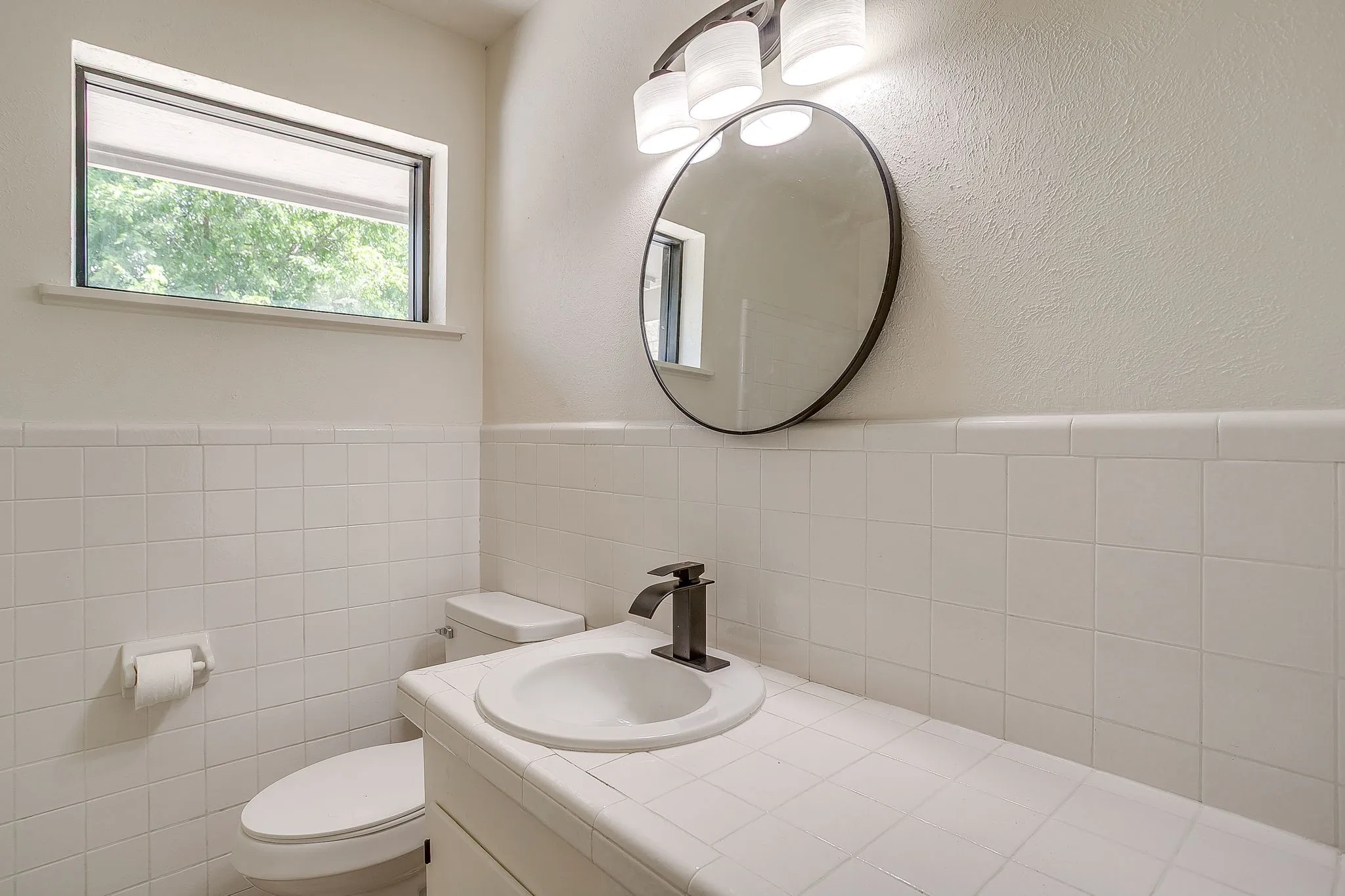 Bathroom featuring white square tile wainscoting, a rectangular window, a circular mirror, a three-light vanity fixture, and a built-in vanity with an integrated sink and black faucet