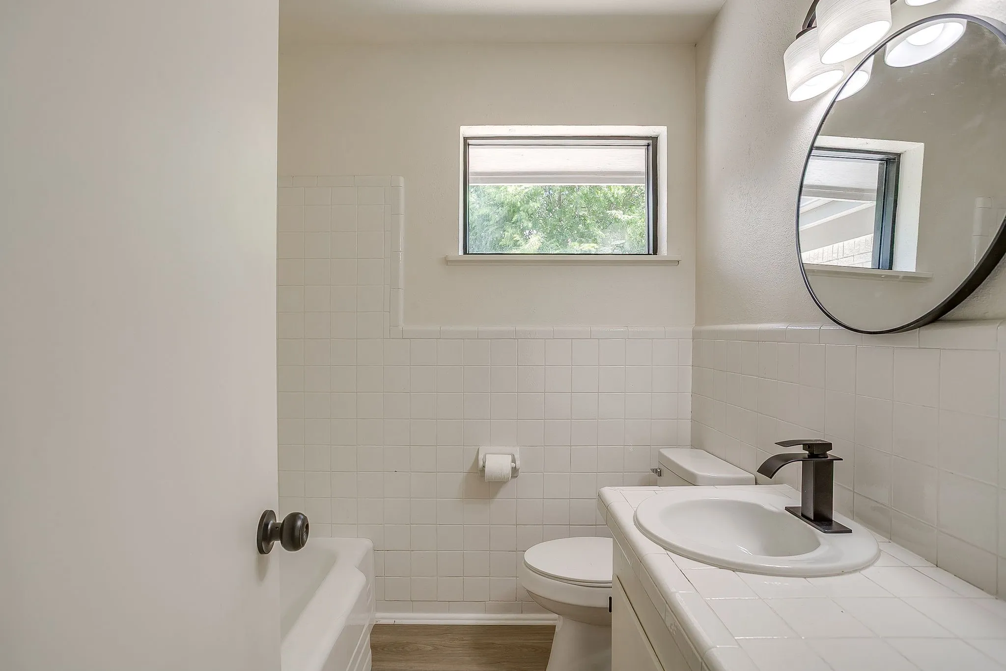 Bathroom featuring white ceramic tile wainscoting, wood-finish flooring, a contemporary black faucet, and a circular wall mirror