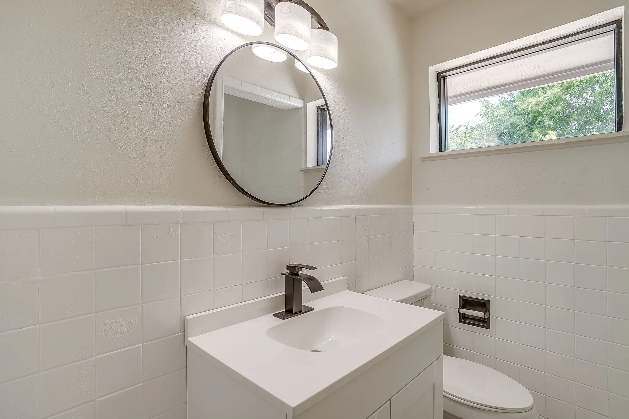 Bathroom featuring a white vanity with integrated sink, black hardware, white subway tile wainscoting, and a round mirror with overhead vanity lighting