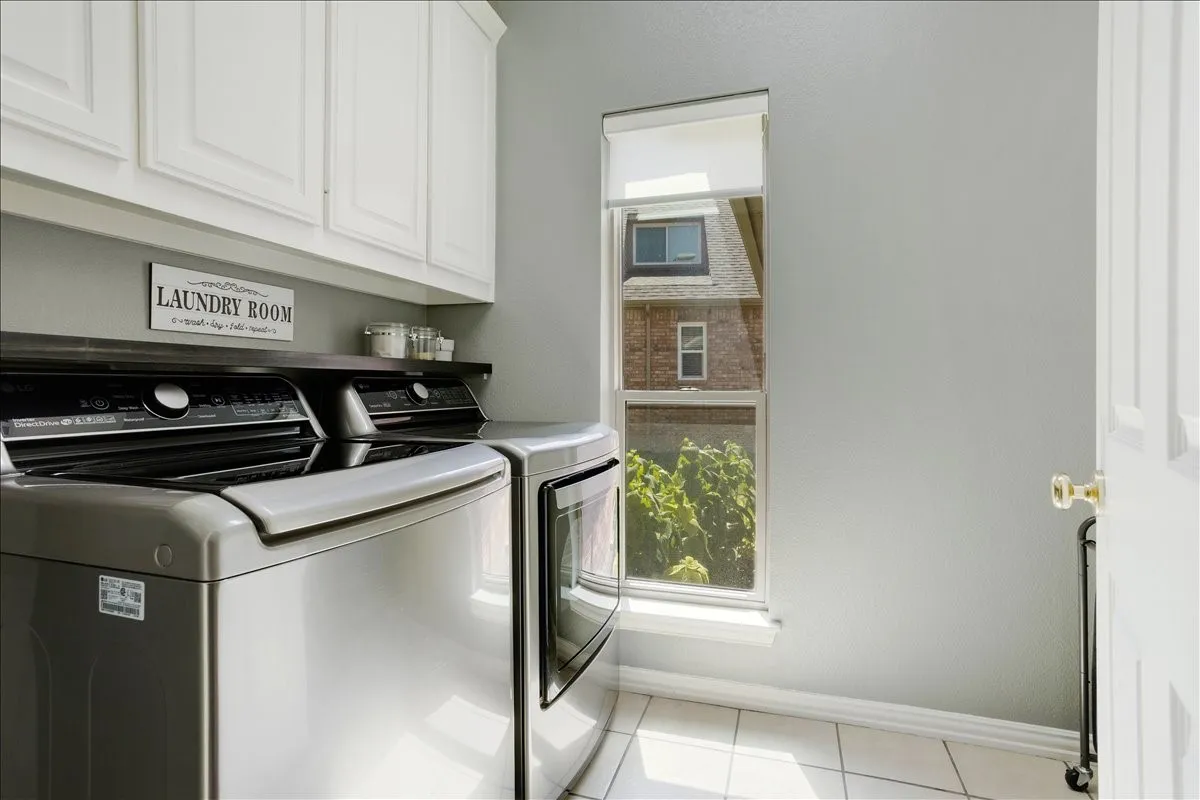 Dedicated laundry area featuring white upper cabinetry, a dark wood-finish countertop, a tall window with roller shade, and white square tile flooring
