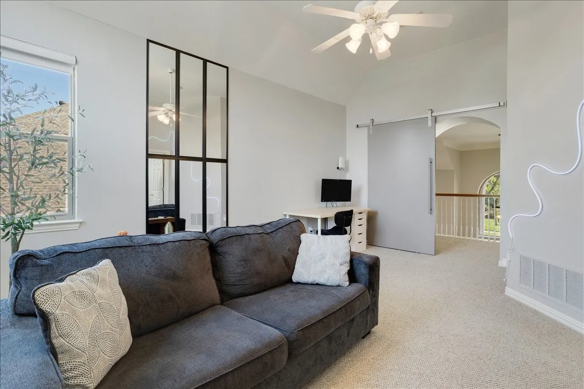Carpeted loft space featuring a vaulted ceiling with a white ceiling fan, a large window, a black-framed glass partition, and a sliding barn door