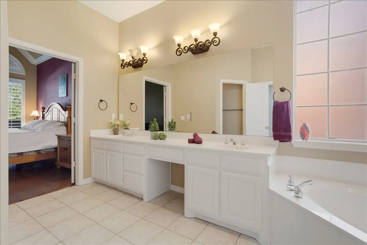 Spacious bathroom featuring an extended dual vanity with white cabinetry, tile flooring, a large soaking tub, and a frosted privacy window