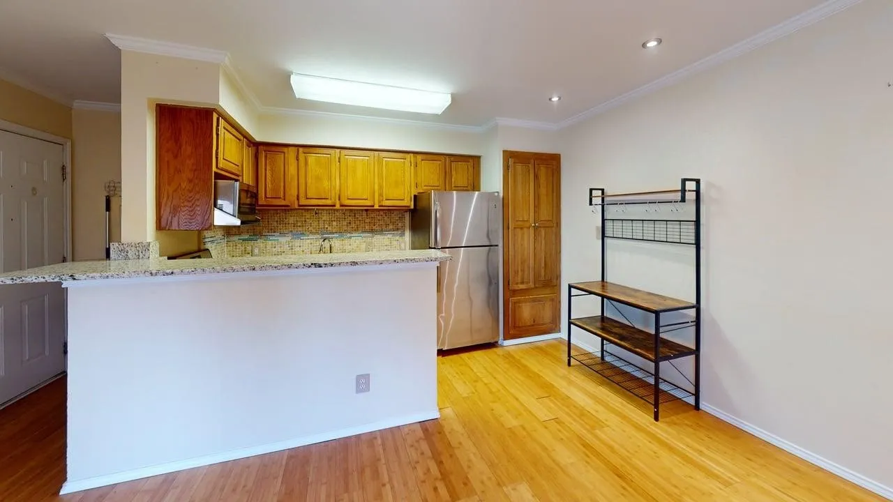 Kitchen area featuring wood-finish flooring, light-colored walls, built-in wooden cabinetry, a stainless steel refrigerator, and a breakfast bar with a light-toned countertop