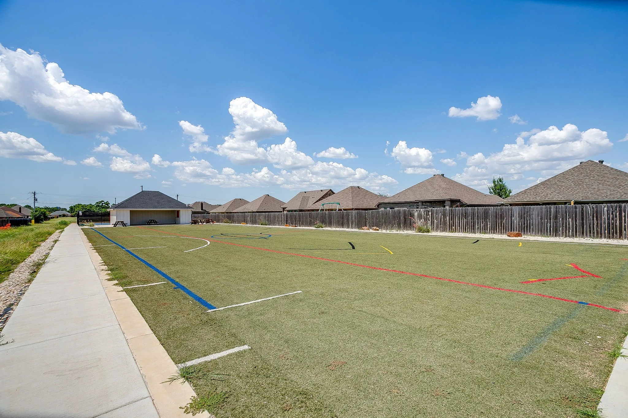 Community turf field with painted game lines, bordered by a concrete sidewalk and a privacy fence
