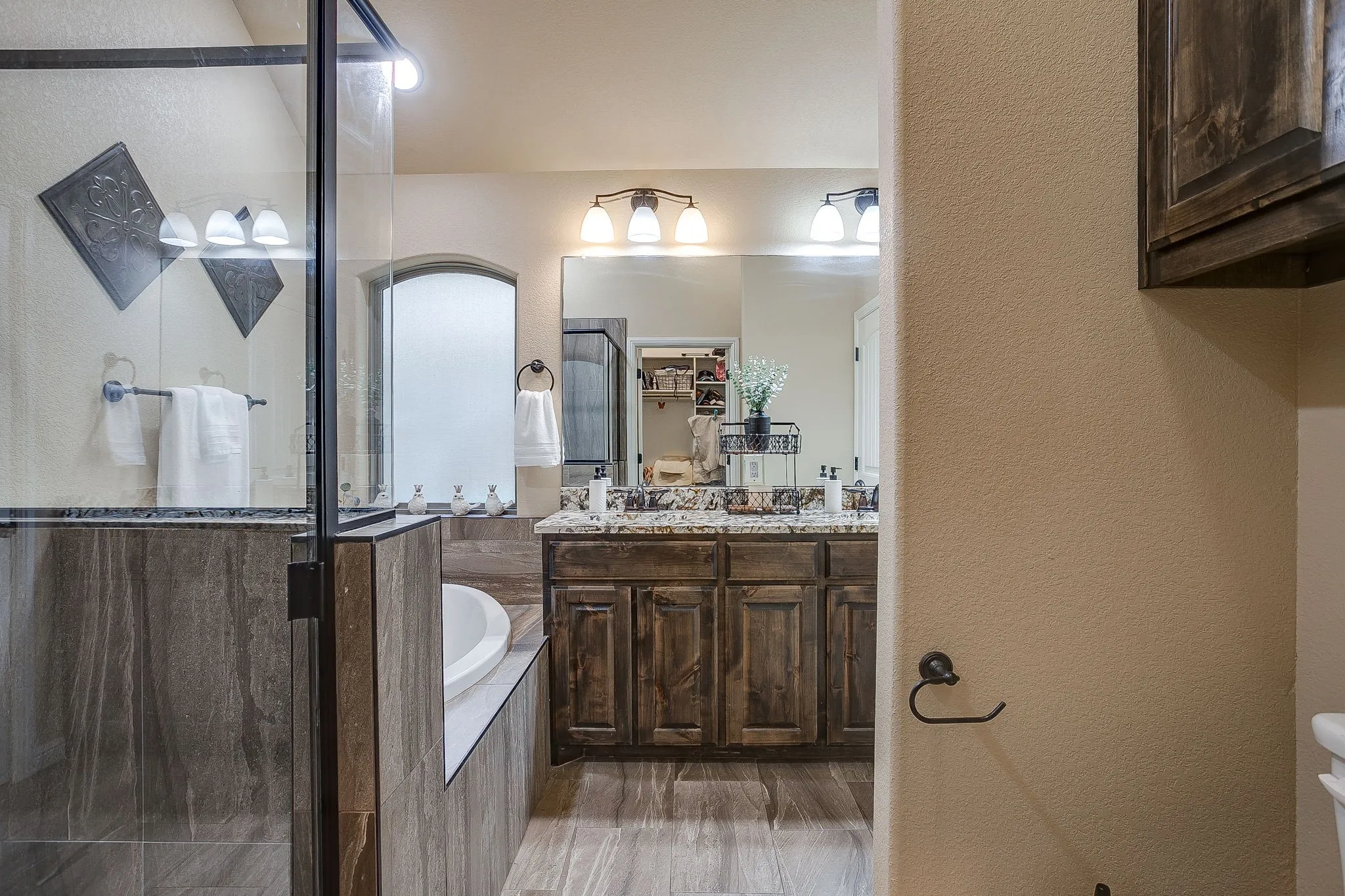 Glass-enclosed shower with dark-toned tile surround, freestanding soaking tub, and a dual vanity featuring wood cabinetry and a light-toned countertop