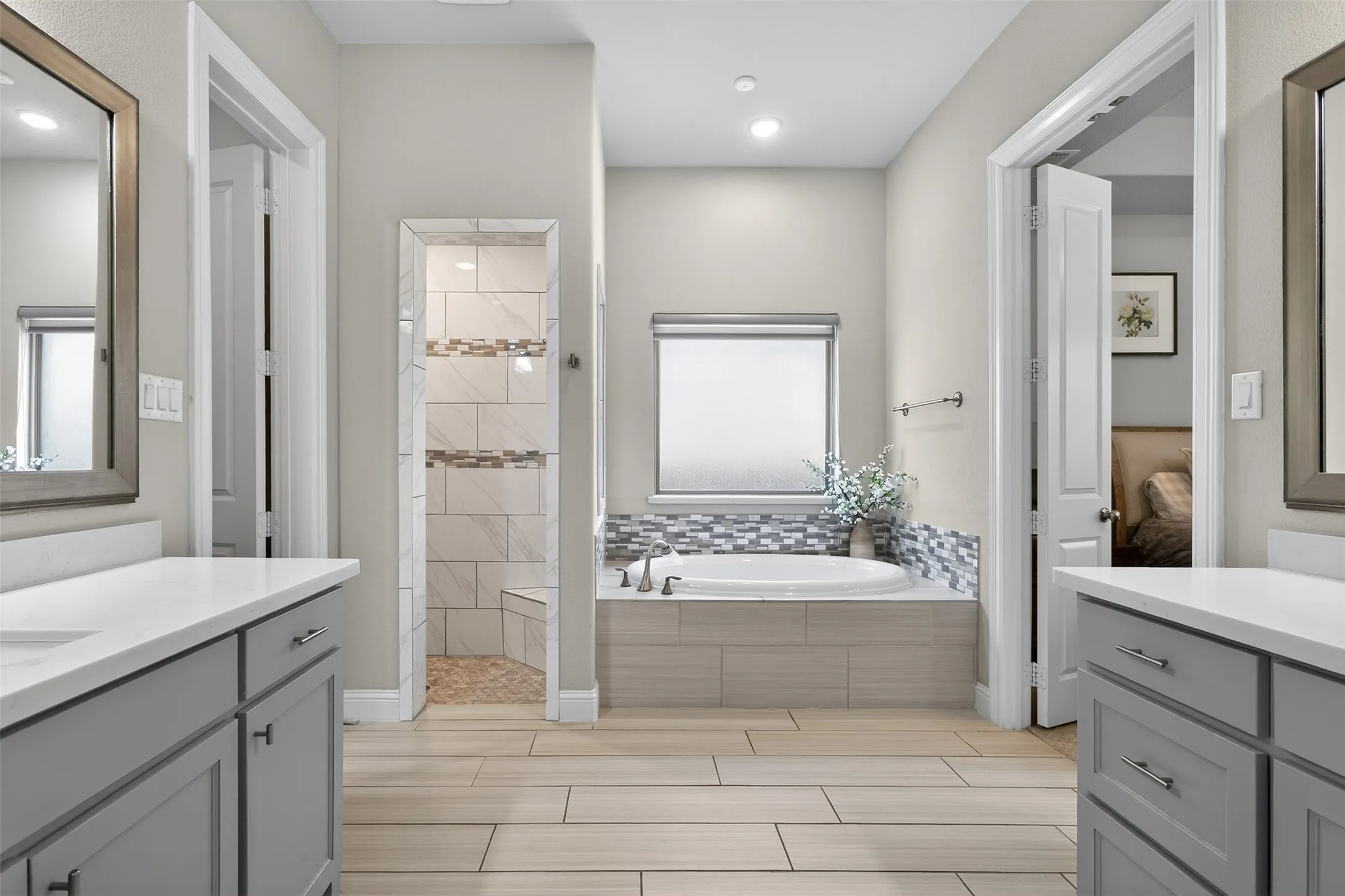 Dual vanity with light gray cabinetry and white countertops, featuring a built-in soaking tub with a mosaic tile surround, a tiled shower with a built-in bench, and wood-finish flooring