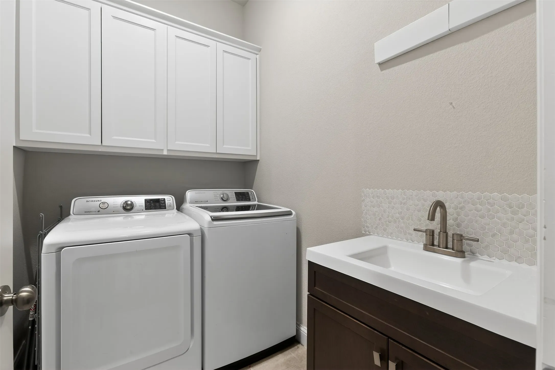 Dedicated laundry area featuring white upper cabinetry, a dark wood-finish vanity with an integrated sink, a brushed nickel faucet, and a scalloped tile backsplash