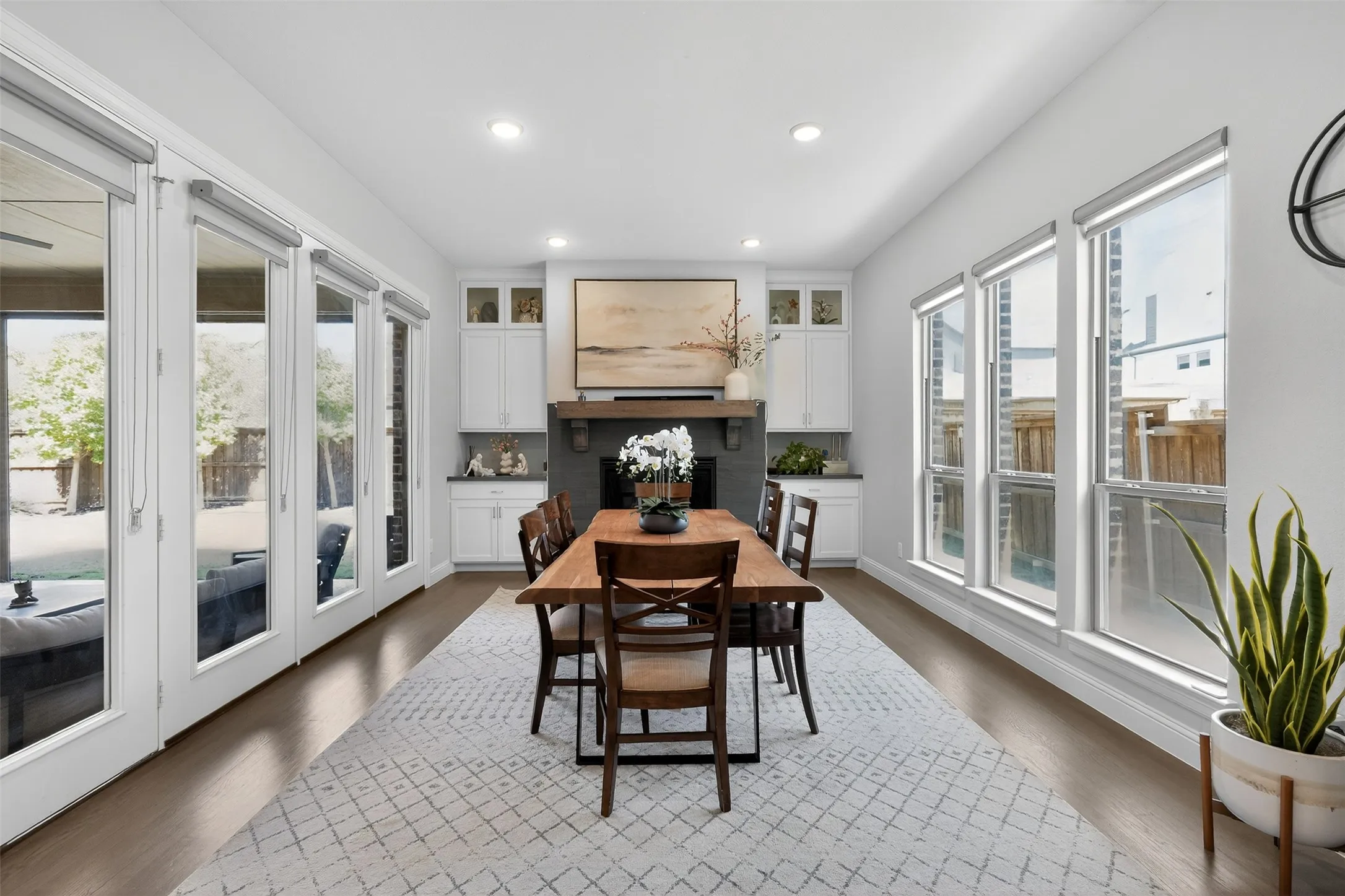 Spacious dining area featuring wood-finish flooring, a fireplace with a dark tile surround and wood mantel, white built-in cabinetry, and multiple large windows and glass-paneled doors offering abundant illumination