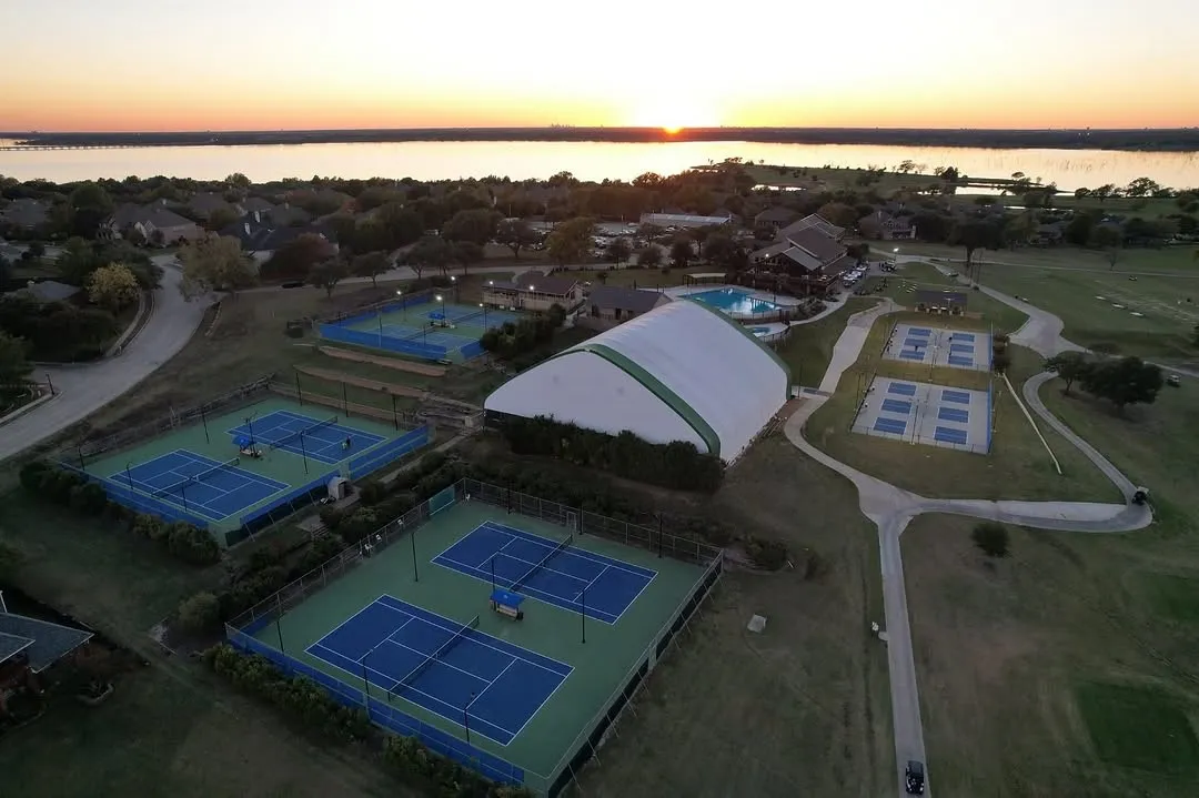 Aerial perspective highlighting multiple outdoor tennis courts, a large indoor court facility, a swimming pool area, and a golf course, all situated alongside a significant body of water