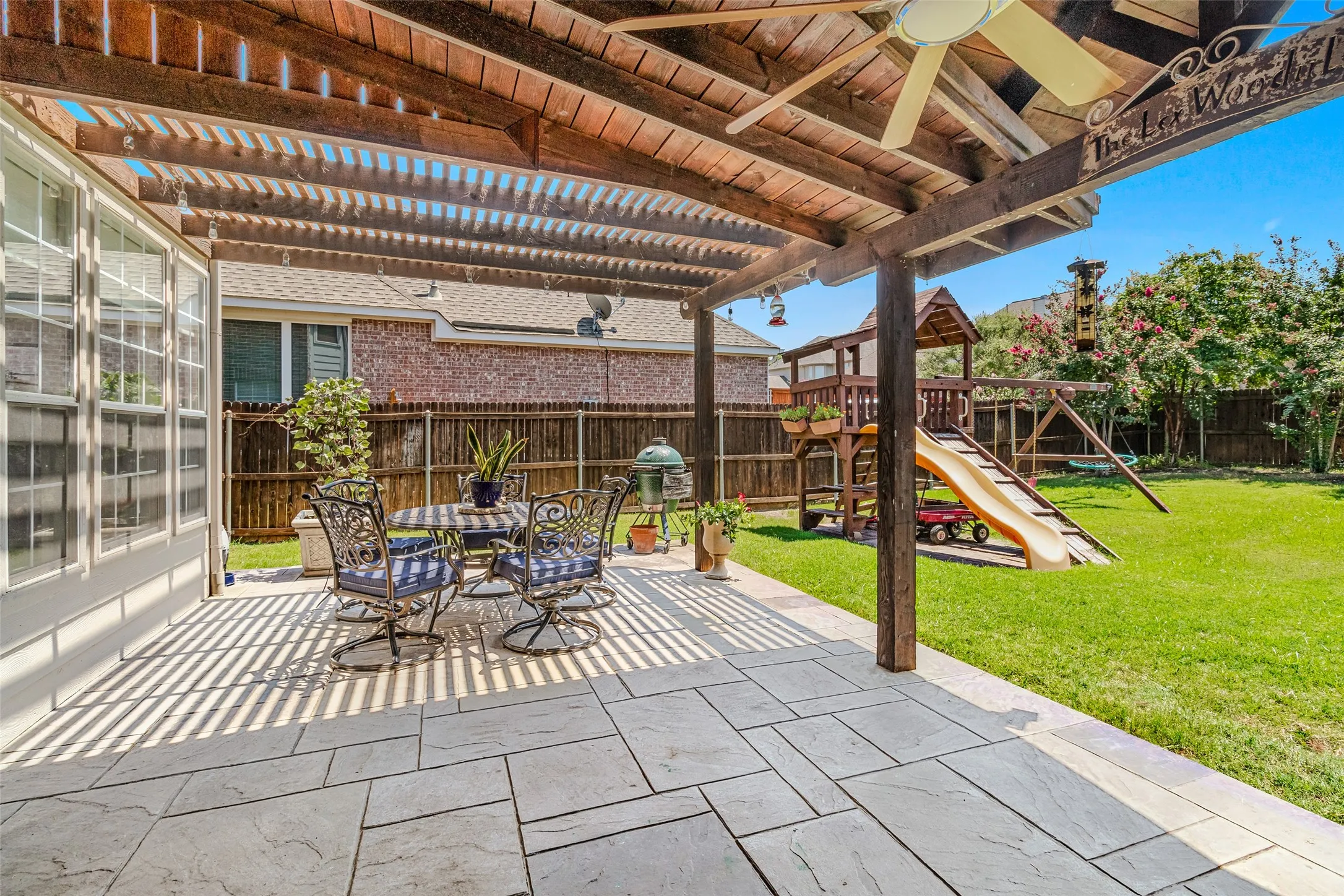 Expansive outdoor patio featuring a wood pergola with a ceiling fan, stone paver flooring, and a large grassy area