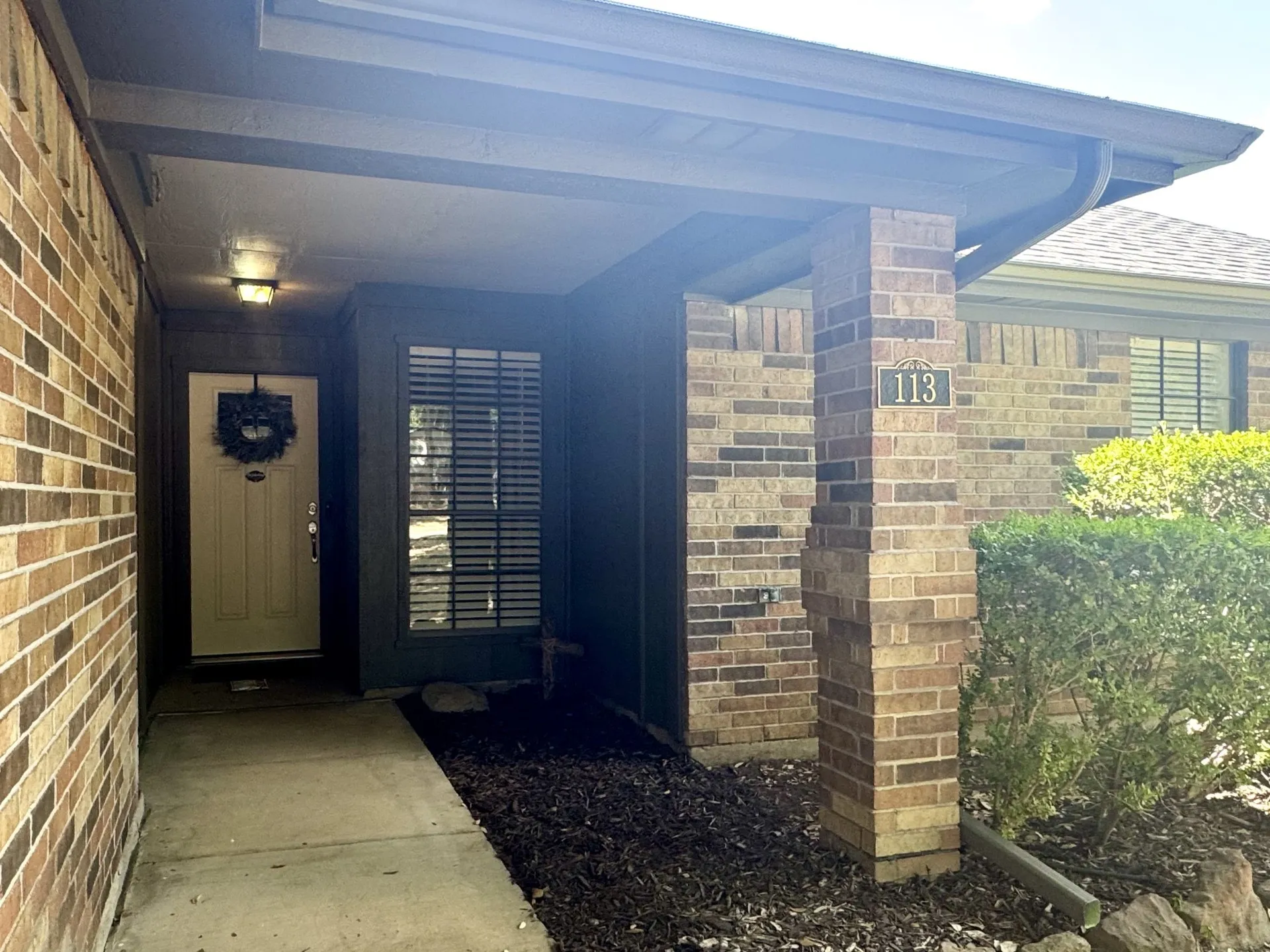 Inviting Covered Front Porch featuring brick column with address marker, rain gutters, an exterior light fixture, and a vertical window with horizontal blinds.