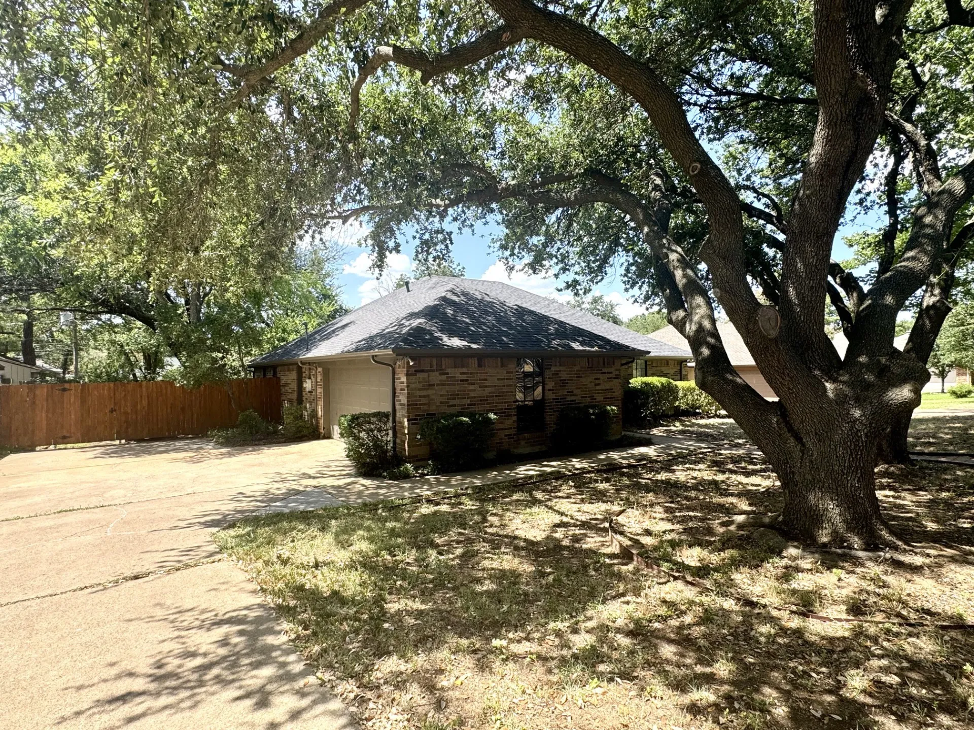 Side-Entry 2-CAR Garage with swing driveway, brick exterior, dark shingle roof, and mature front yard trees. Newer WOOD Fence too!