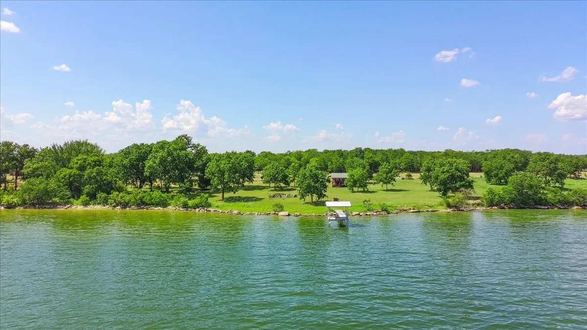 Expansive waterfront property featuring a private boat dock, lush green lawn, mature trees, and a distant structure