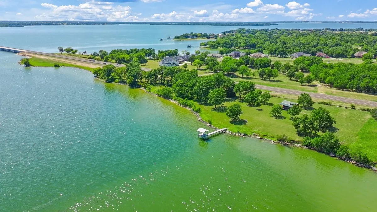 Waterfront property featuring a private boat dock, expansive green lawn, mature trees, and a concrete-paved road