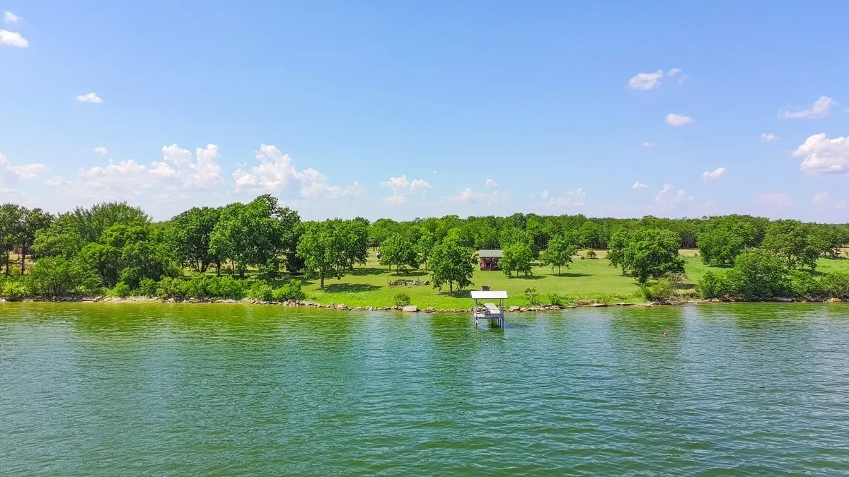 Expansive waterfront property featuring a private boat dock, lush green lawn, mature trees, and a distant cabin