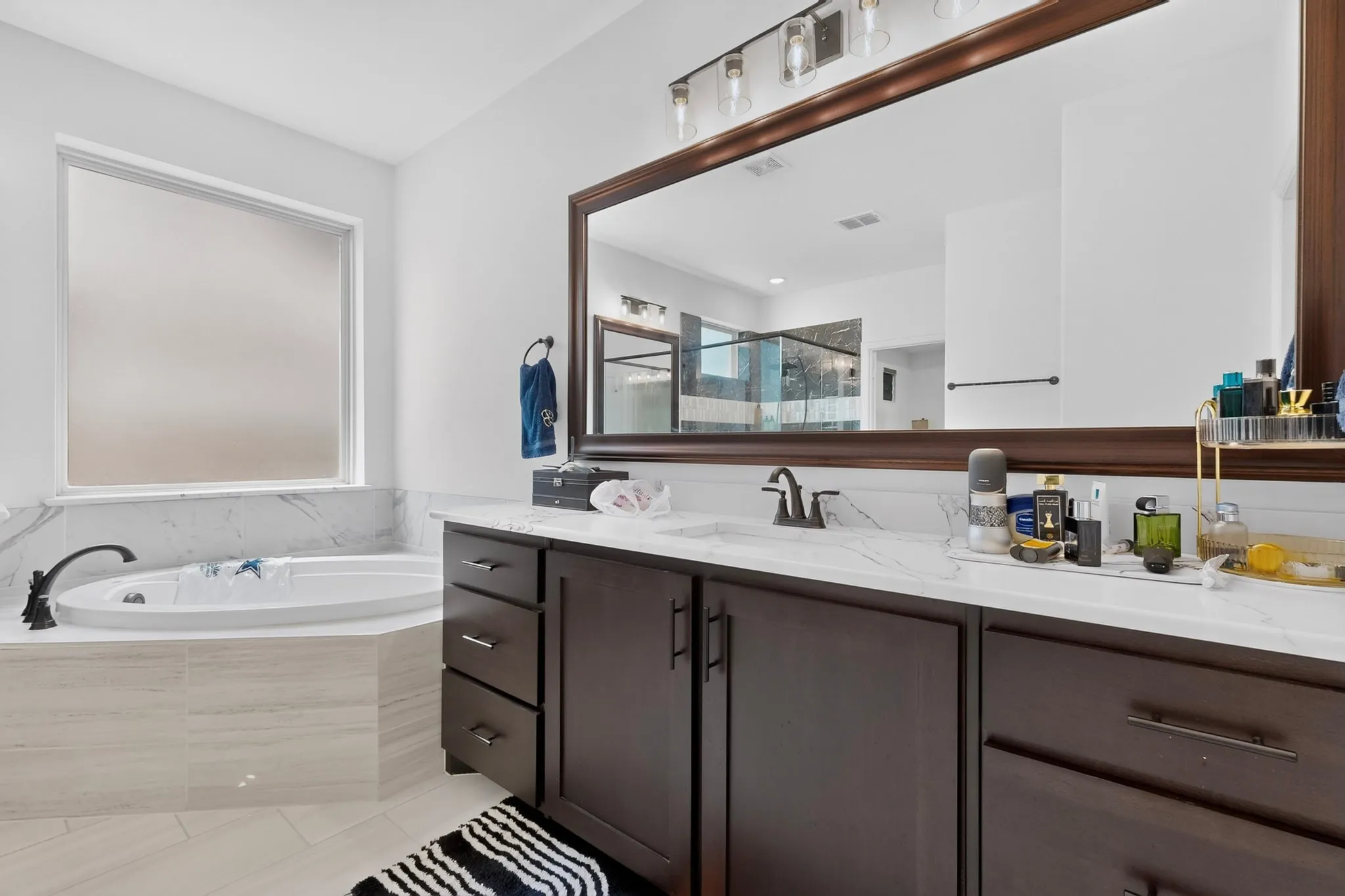 Bathroom featuring a deep soaking tub with surrounding tile, a large vanity with dark wood-finish cabinetry, a single basin sink, and a spacious framed mirror