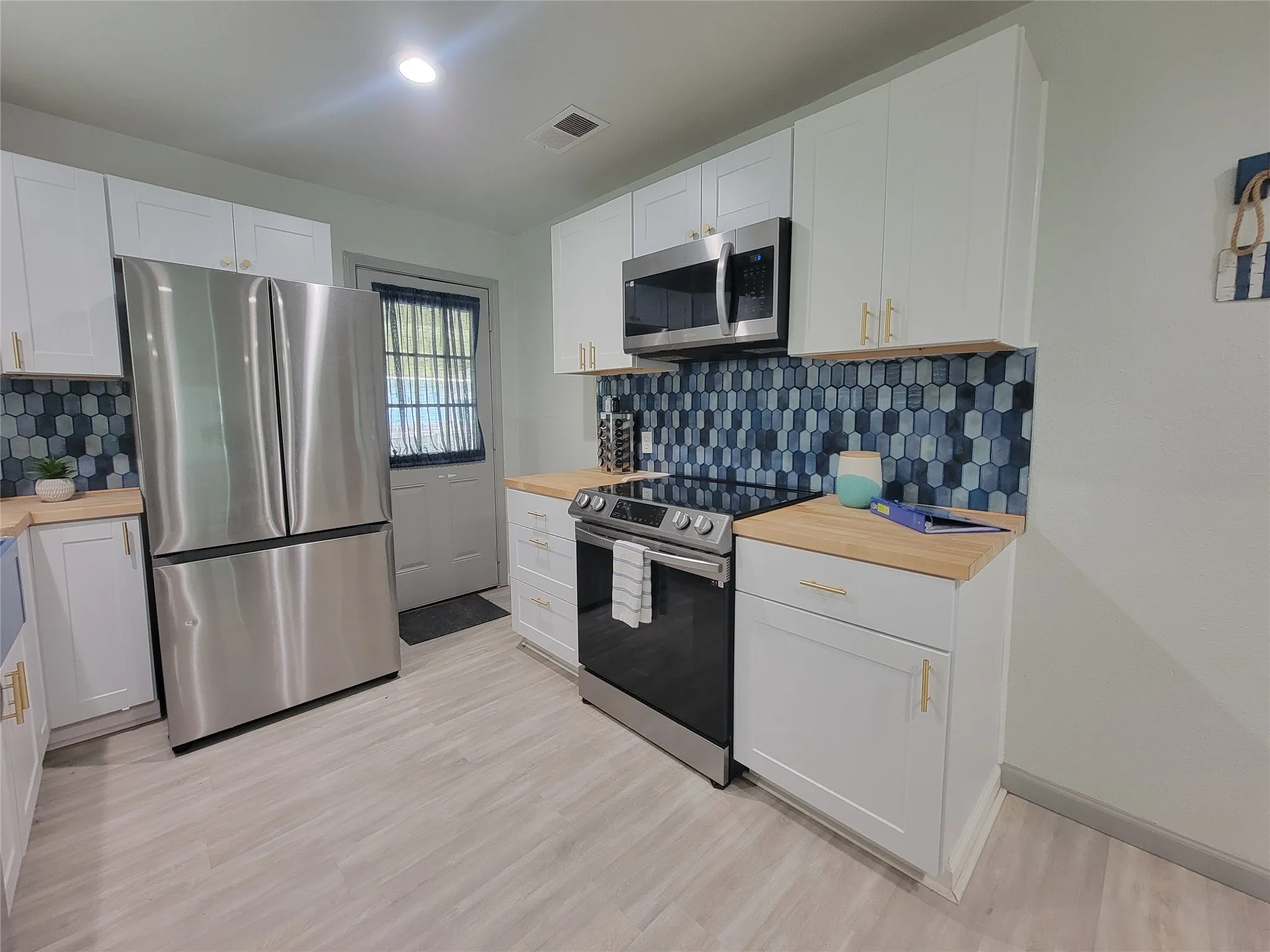 Modern kitchen featuring stainless steel appliances, light wood-finish countertops, white cabinetry with gold-tone hardware, a blue and gray geometric tile backsplash, and light wood-finish flooring