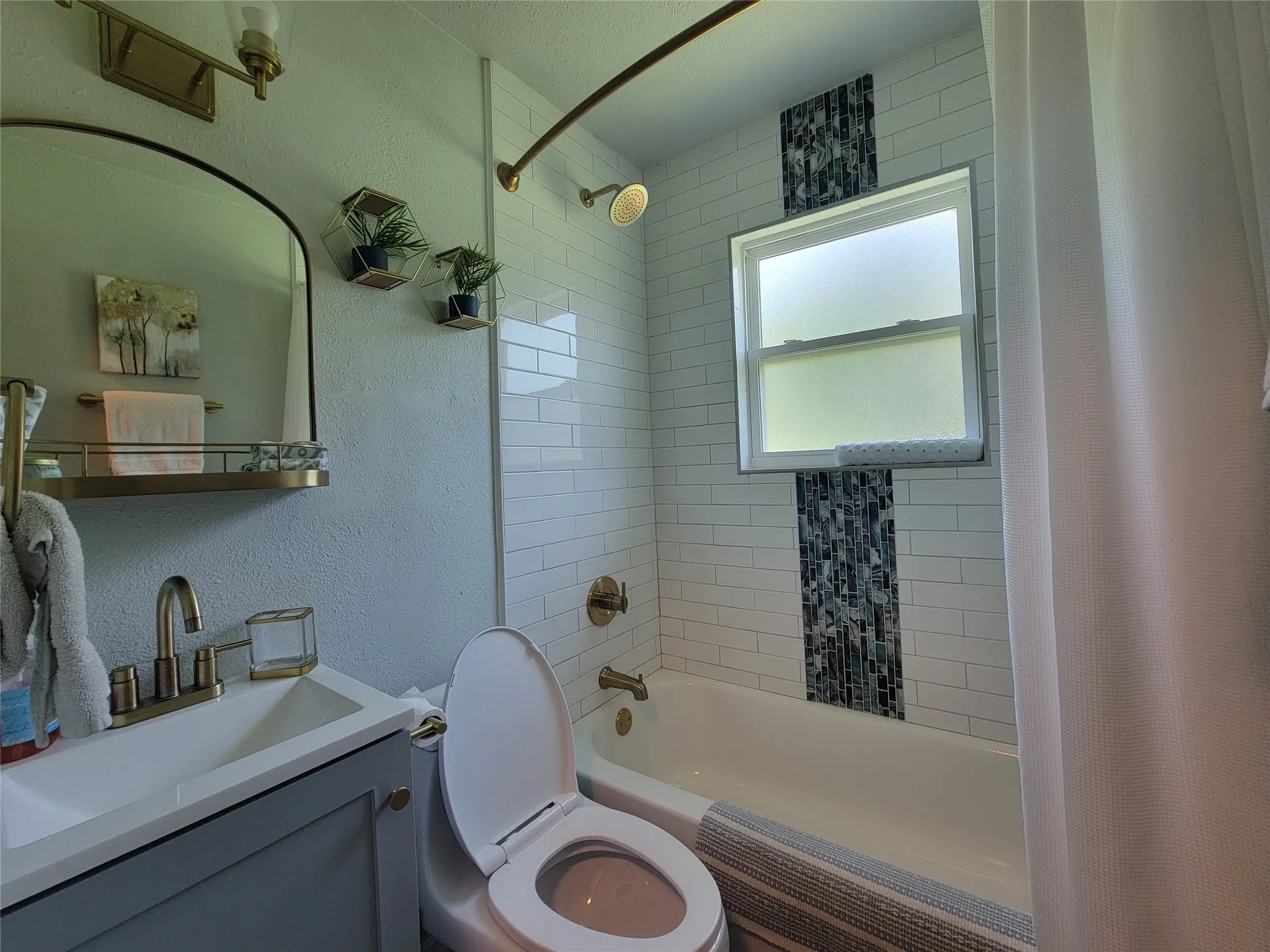 Bathroom featuring a white subway tile shower surround with a mosaic tile vertical accent, a window, a built-in tub, a vanity with a white countertop and a brass-finish faucet, and an arched mirror