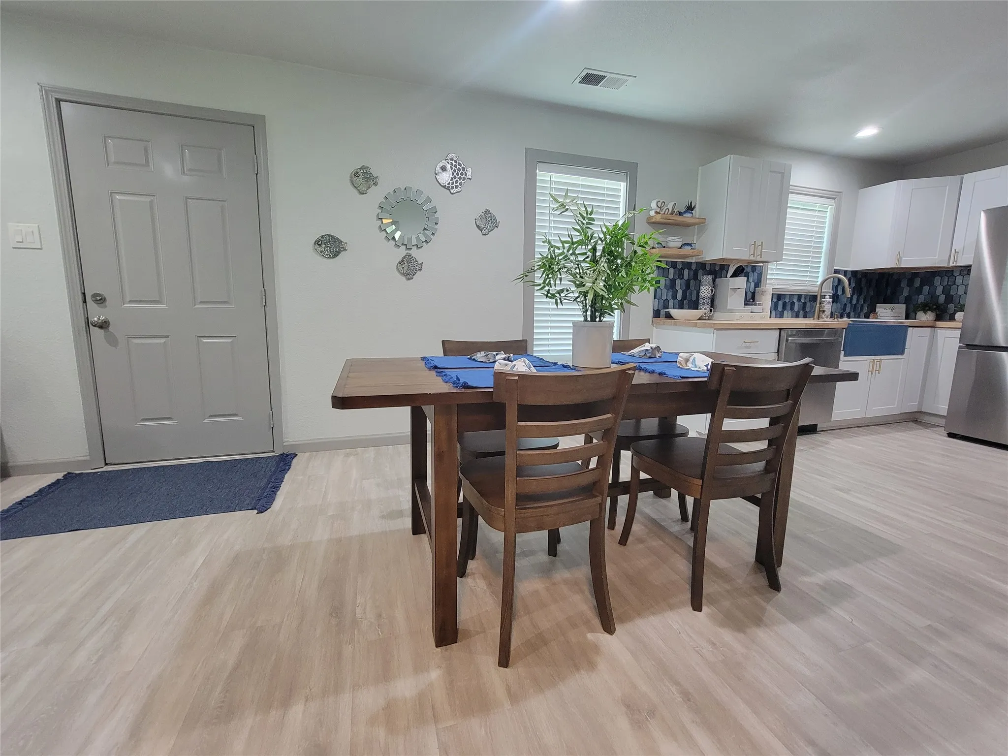 Open-concept living space featuring light wood-finish flooring, a contemporary kitchen with white cabinetry and a blue tile backsplash, stainless steel appliances, and a farmhouse-style sink