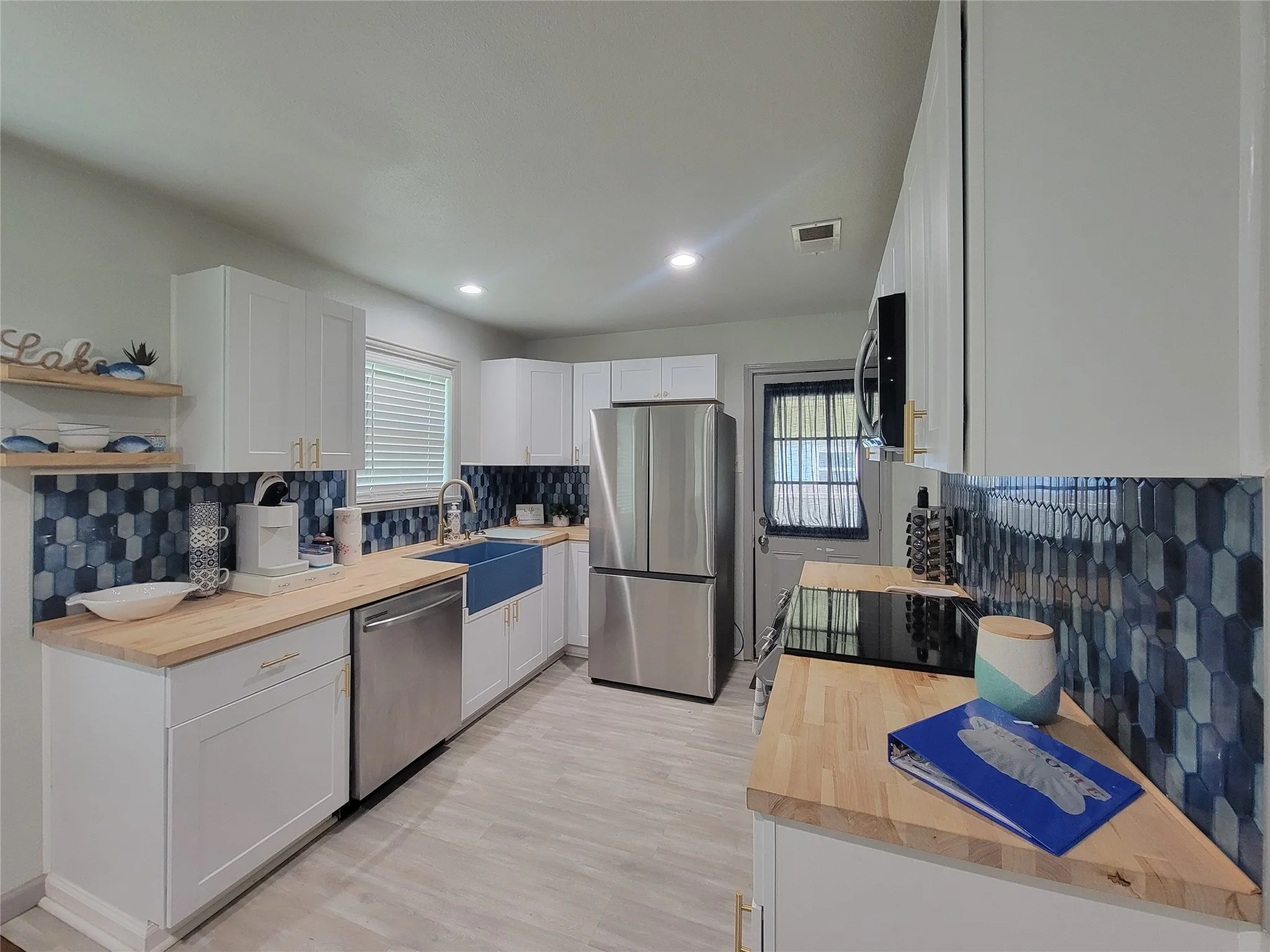 Bright kitchen featuring white cabinetry with gold-tone hardware, a blue hexagonal tile backsplash, wood-finish countertops, and stainless steel appliances including a refrigerator and dishwasher