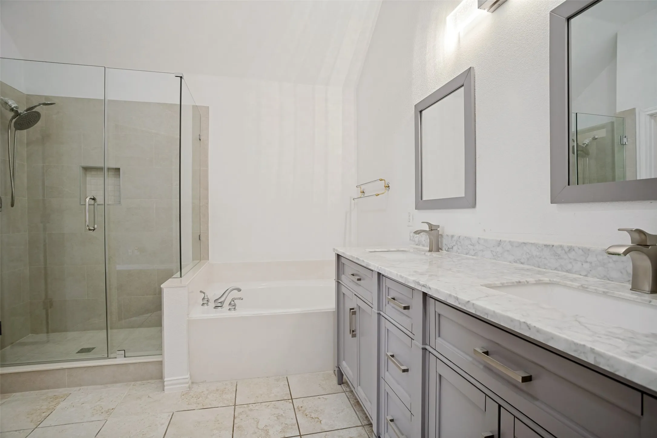 Bathroom featuring a dual vanity with grey cabinetry and light-toned countertops, a glass-enclosed shower with tiled walls and a built-in niche, a white soaking tub, and square floor tiles