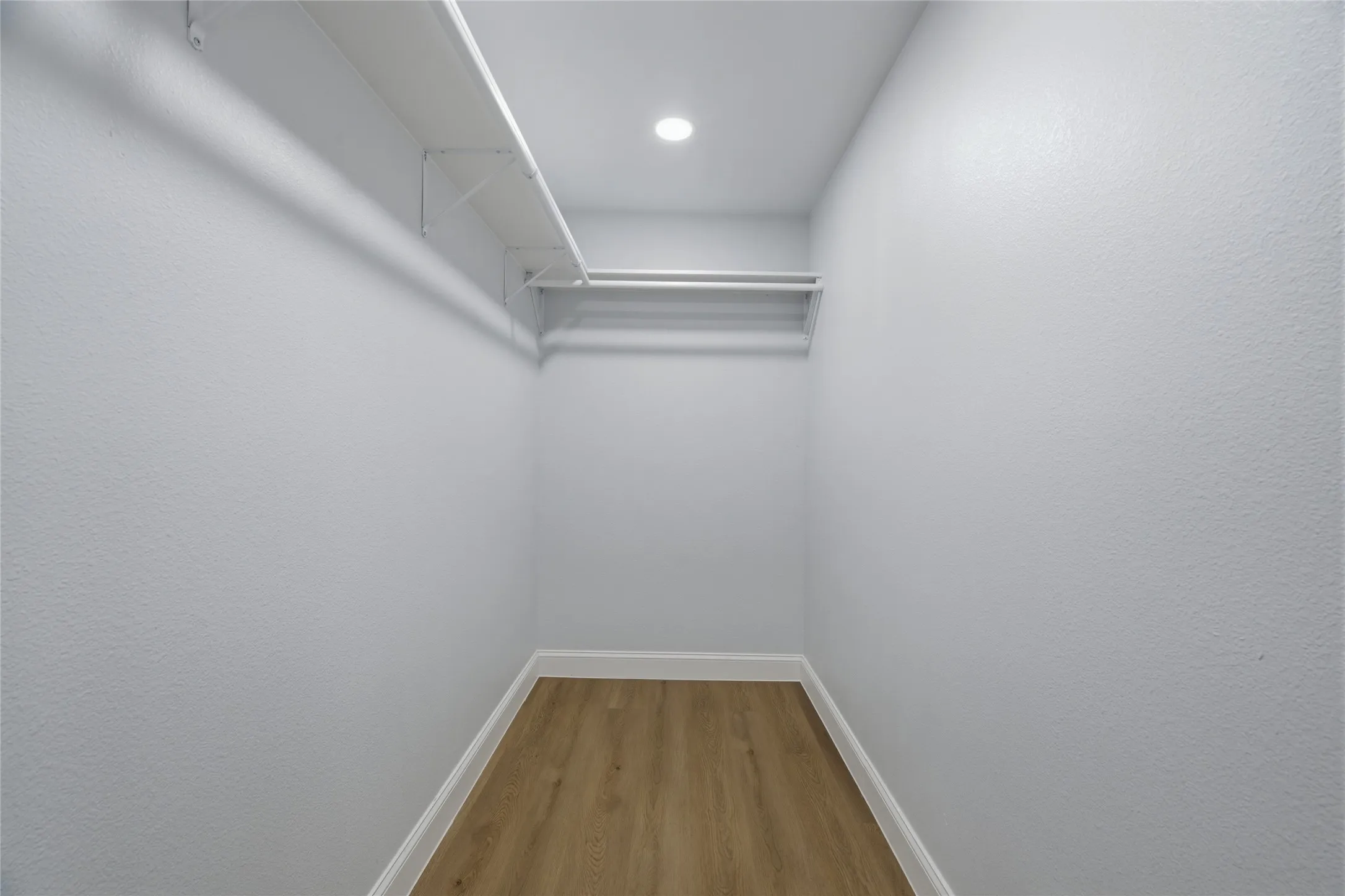 Walk-in closet featuring light wood-finish flooring, white baseboards, a single recessed ceiling light, and built-in white shelving with a hanging rod