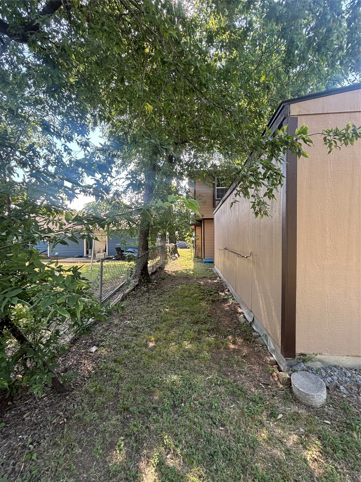 Expansive side yard featuring a chain-link fence, mature trees, and a beige exterior structure with brown trim