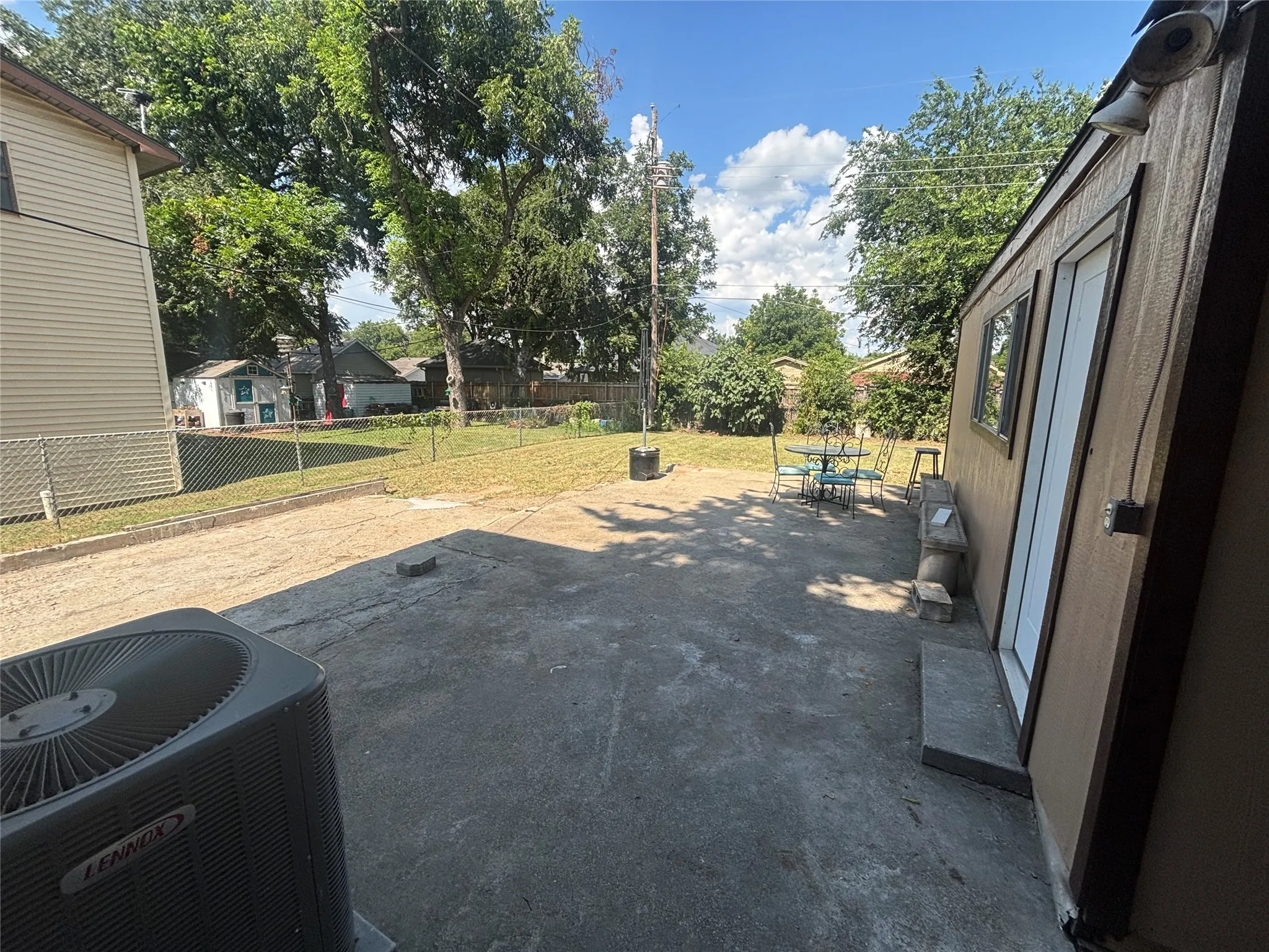 Spacious concrete patio area featuring a Lennox HVAC unit, a separate exterior structure with a window and door, and a chain-link fence bordering a grassy area with mature trees