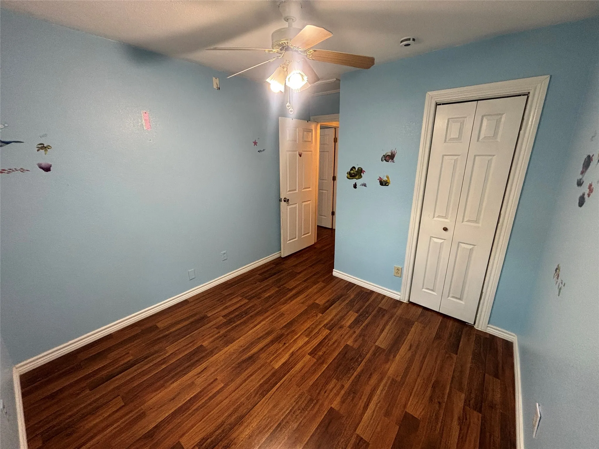 Interior room featuring wood-finish flooring, white baseboards, a ceiling fan with light fixture, and a white bi-fold closet door