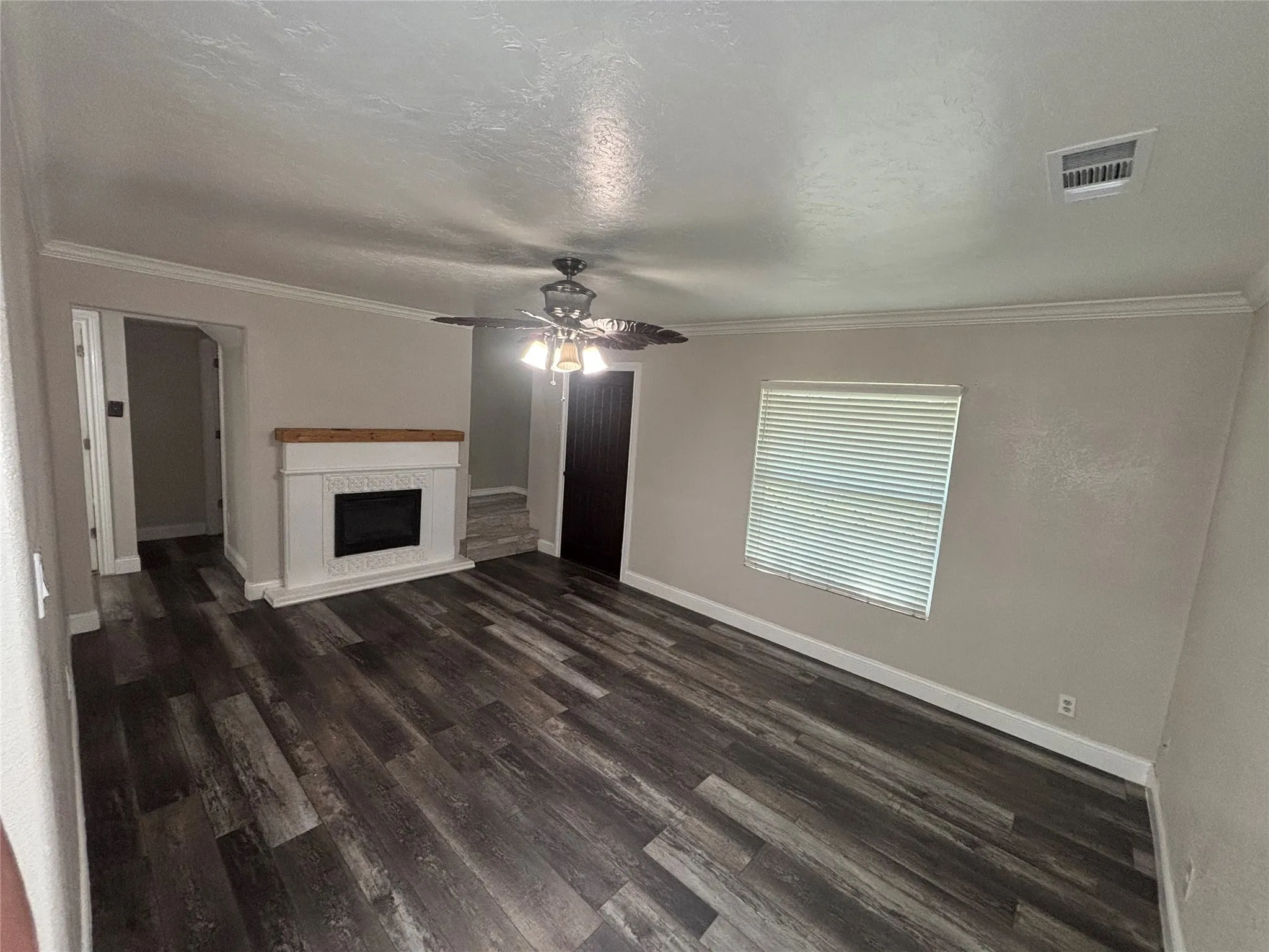 Living area featuring wood-finish flooring, a white mantel fireplace with a wood top, a ceiling fan, crown molding, and a window with blinds