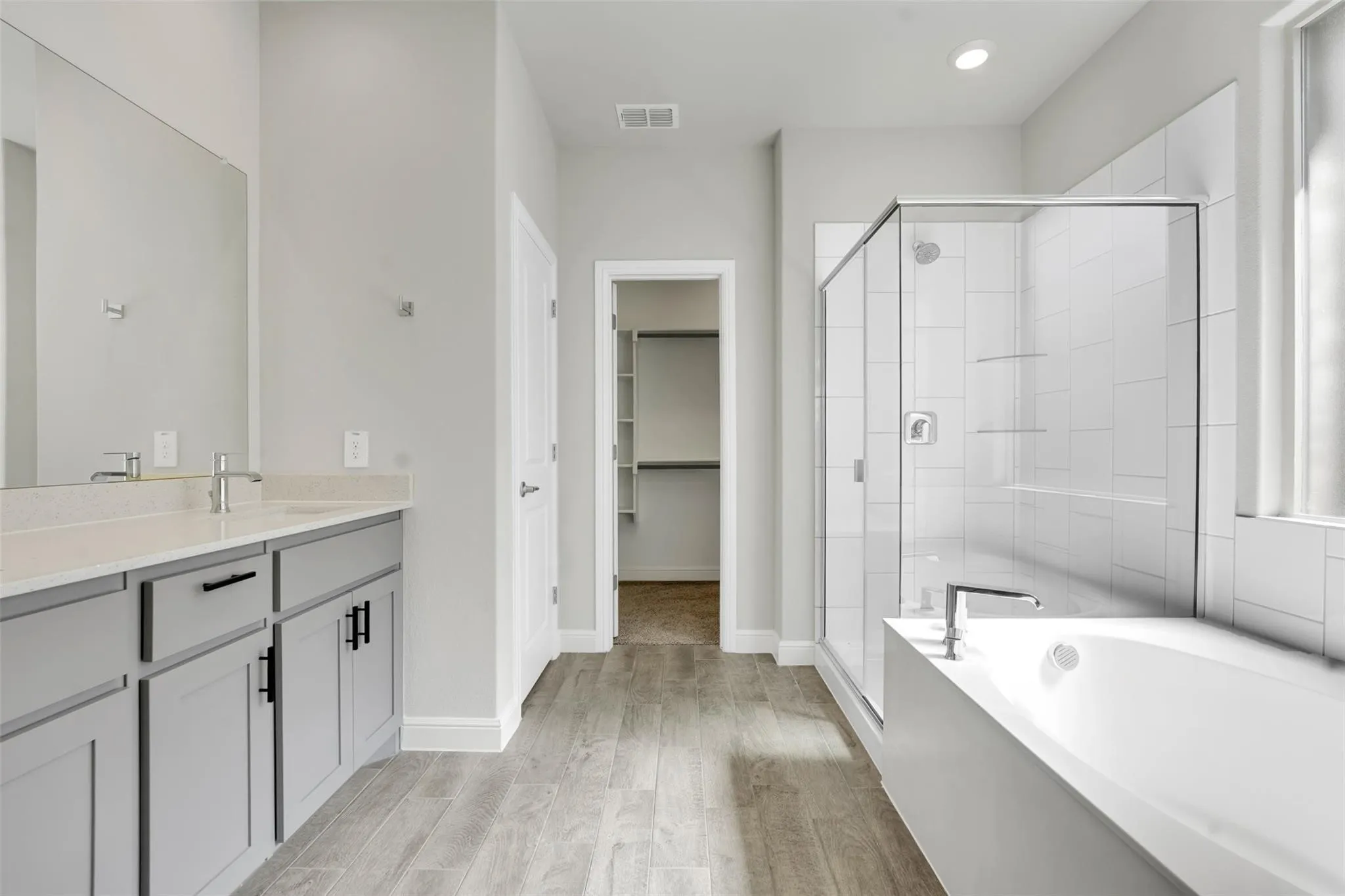 Bathroom featuring dual vanity with gray cabinetry, a walk-in shower with clear glass enclosure, a built-in bathtub, and wood-finish flooring