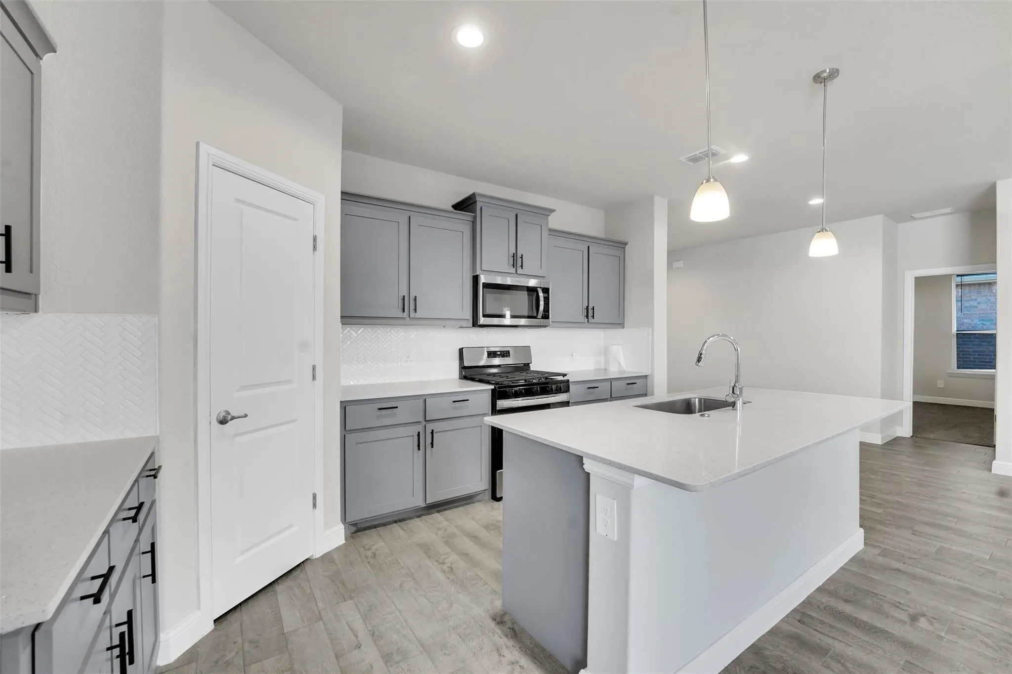 Modern kitchen featuring gray cabinetry, stainless steel appliances, a herringbone tile backsplash, an island with an undermount sink, and wood-finish flooring