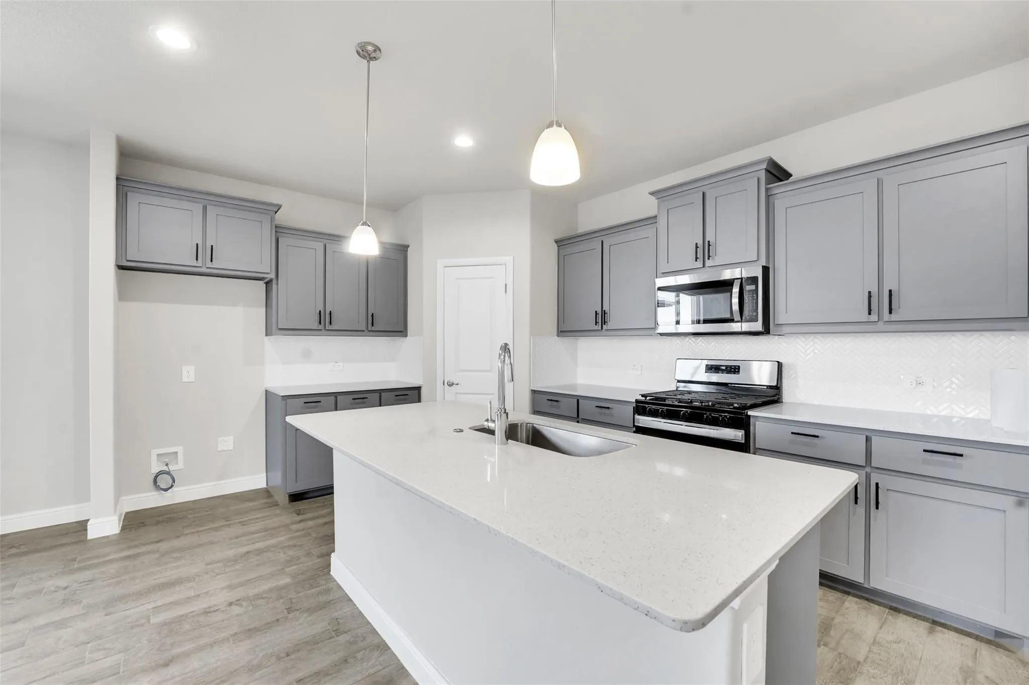Contemporary kitchen featuring a spacious island with an undermount sink, light-toned countertops, gray cabinetry, stainless steel appliances, and wood-finish flooring