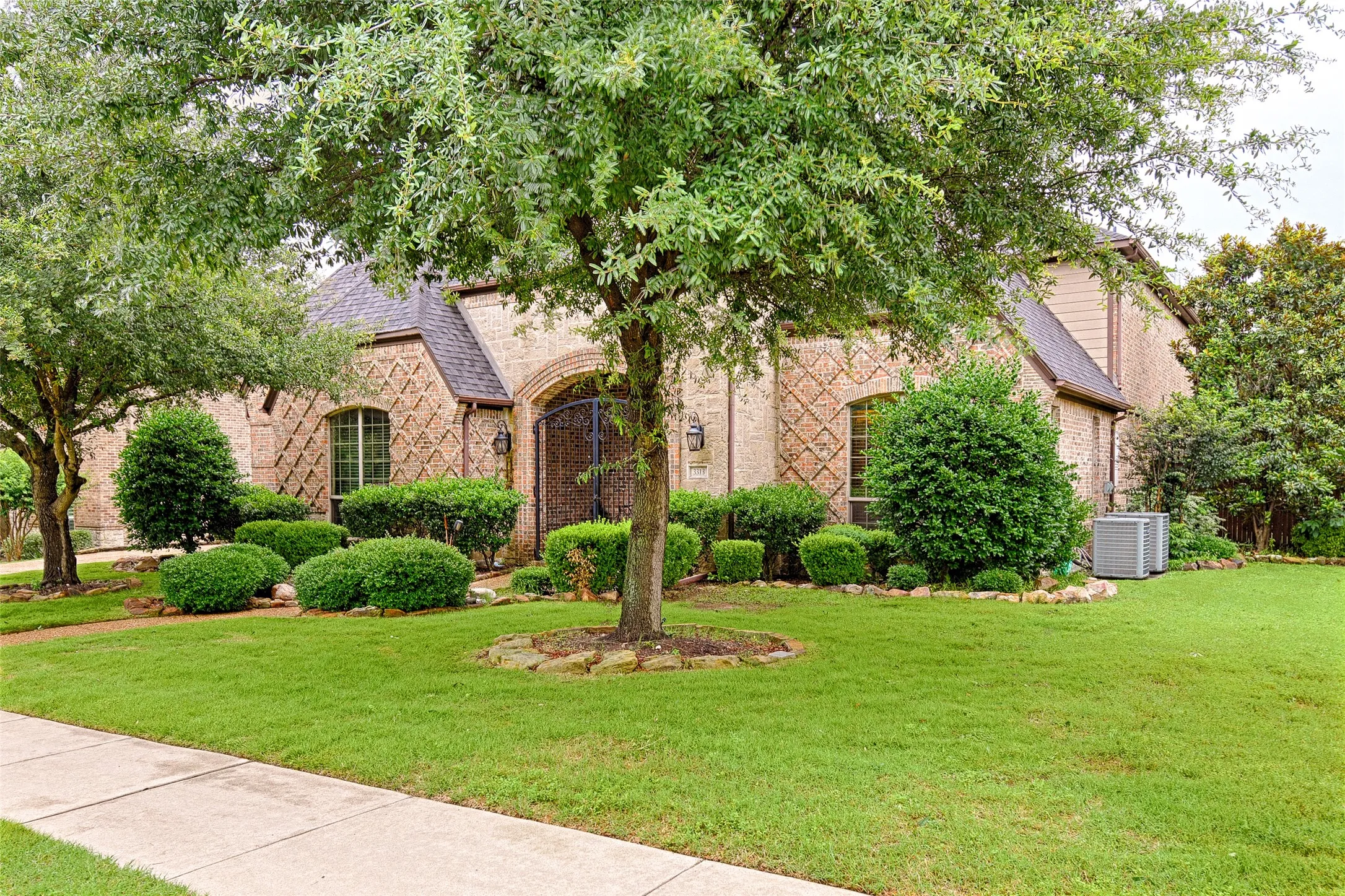 Brick exterior featuring decorative diamond-patterned masonry, arched entryway with wrought iron gate, and mature trees providing natural shading