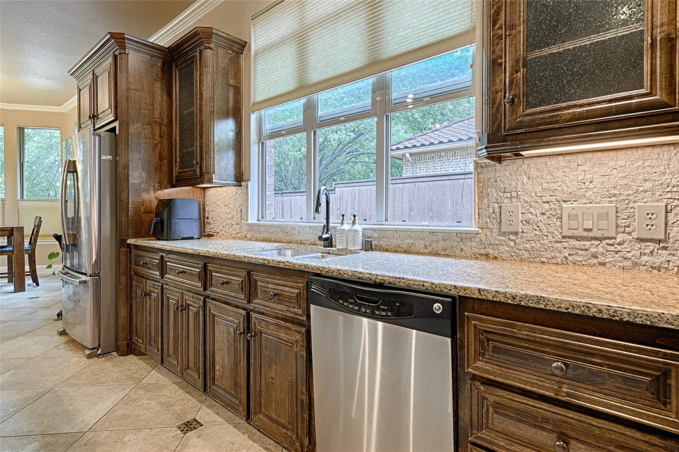 Kitchen featuring custom wood cabinetry, granite countertops, a textured tile backsplash, a stainless steel dishwasher, and a large window above the sink