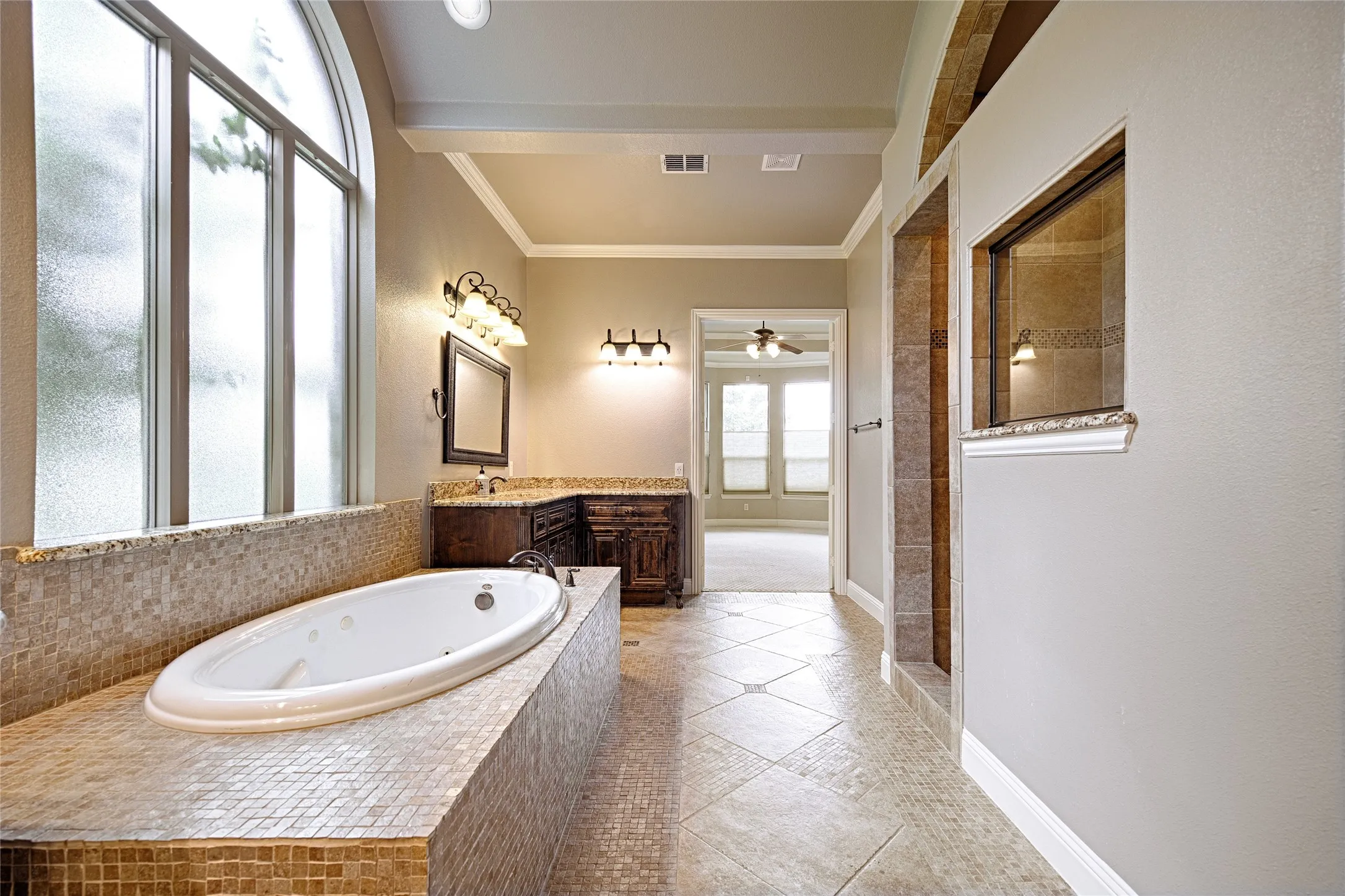 Soaking tub with mosaic tile surround, dual vanity with dark wood cabinetry and granite-style countertops, arched window, and tiled flooring