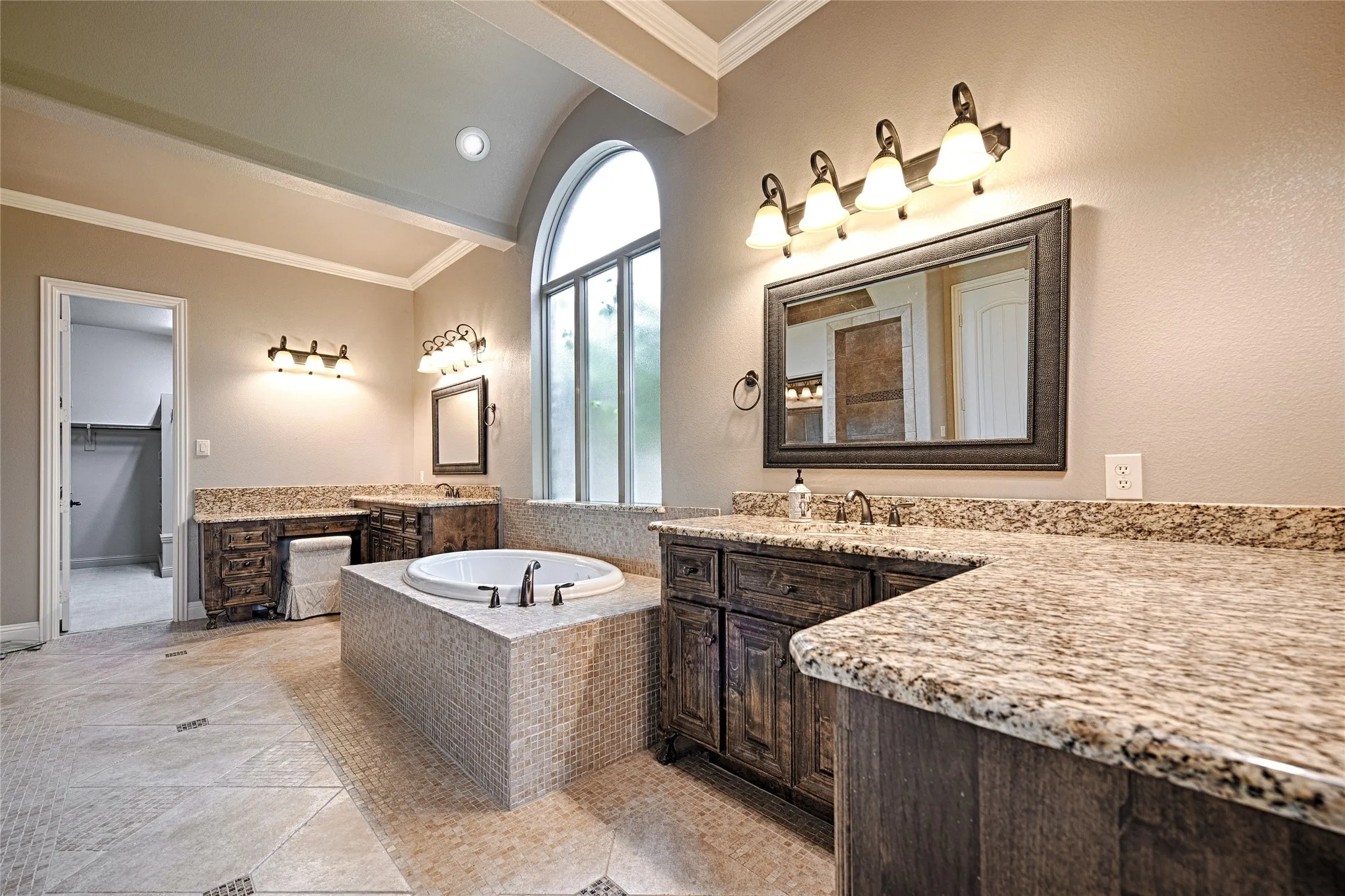Bathroom featuring a built-in tub with mosaic tile surround, arched window, dual vanities with light-toned countertops and dark wood cabinetry, and tiled flooring