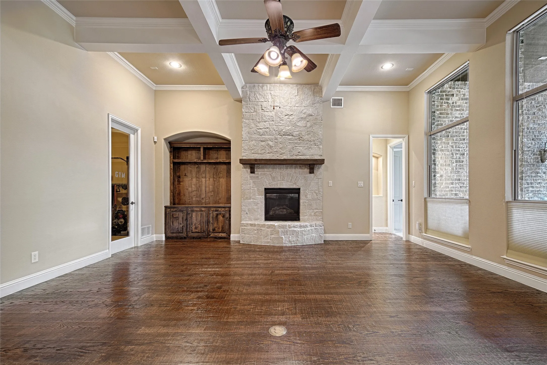 Spacious living area featuring coffered ceilings, a stone fireplace with a wood mantle, wood-finish flooring, and large windows with blinds