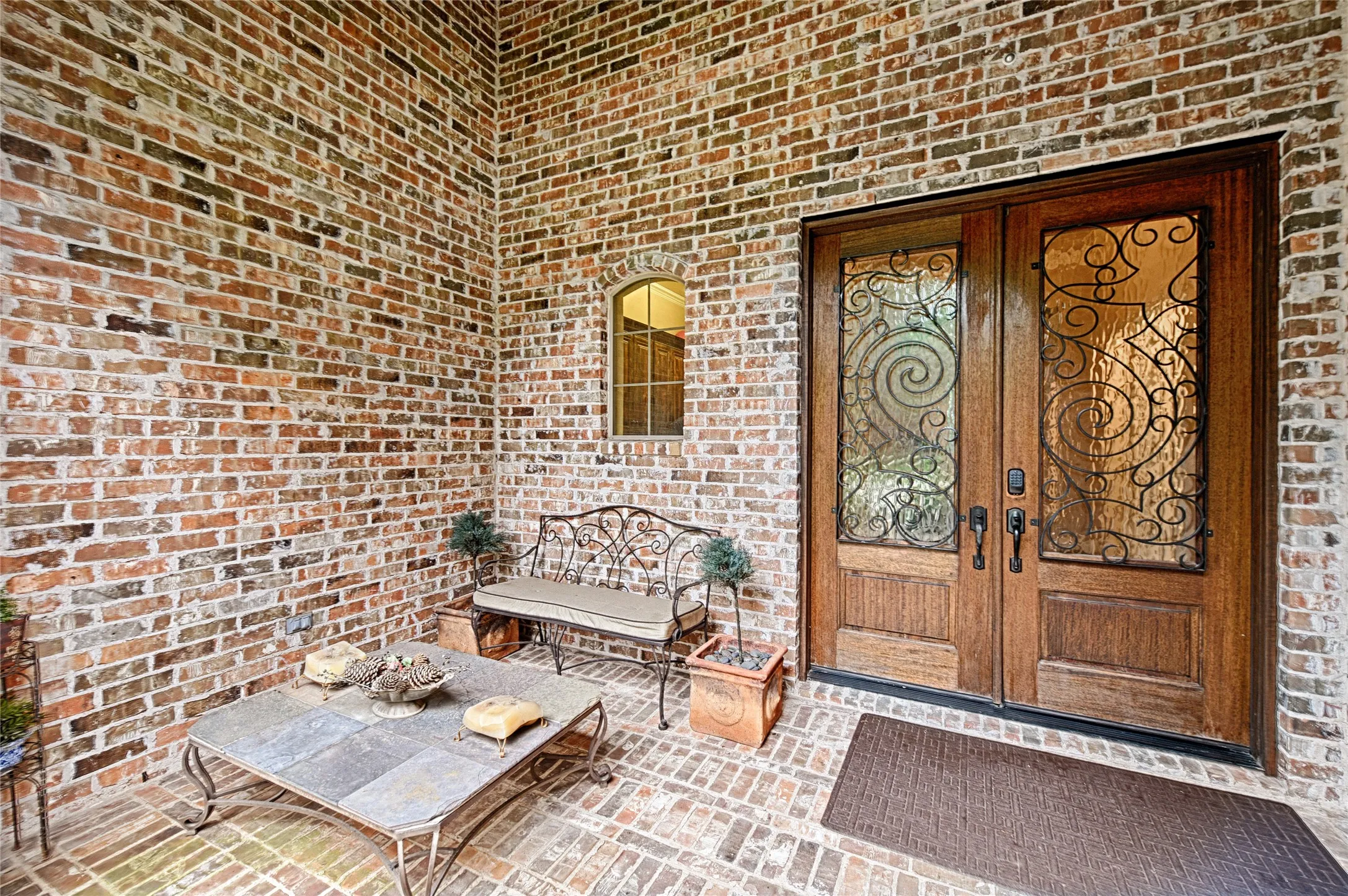 Brick facade entry featuring double wooden doors with decorative ironwork and obscured glass panels, a brick paver porch, and an arched window